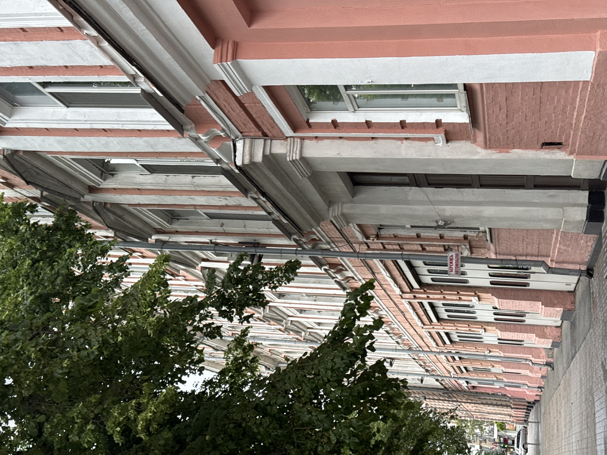A long, multi-story building facade, located in Kyiv, Ukraine, is viewed from an upward-angled, street-level perspective on a daytime, possibly overcast, day. The building features a distinctive salmon-pink color on its main walls, contrasted by white architectural trim, cornices, and ledges. These elements create a repetitive pattern along the facade, highlighting numerous rectangular windows, some of which are slightly ajar.
Below the windows, concrete support beams and decorative brickwork add to the building's historical character. Utility elements such as grey drainpipes or electrical conduits are visible running along the building's surface. Lush green trees occupy the left and lower-left foreground, their dense foliage partially obscuring the lower sections of the building. To the right, a textured grey sidewalk extends into the distance, where the vague outlines of distant urban structures and a white vehicle can be seen.
A prominent white rectangular sign with a red border and red Cyrillic text is affixed to the building's upper facade. The text reads "БЕРЕЖИСЬ АВТОМОБІЛЯ!", which translates to "BEWARE OF THE CAR!" or "CAUTION: VEHICLE!". No individuals are present, and the scene is static, depicting a quiet urban street.