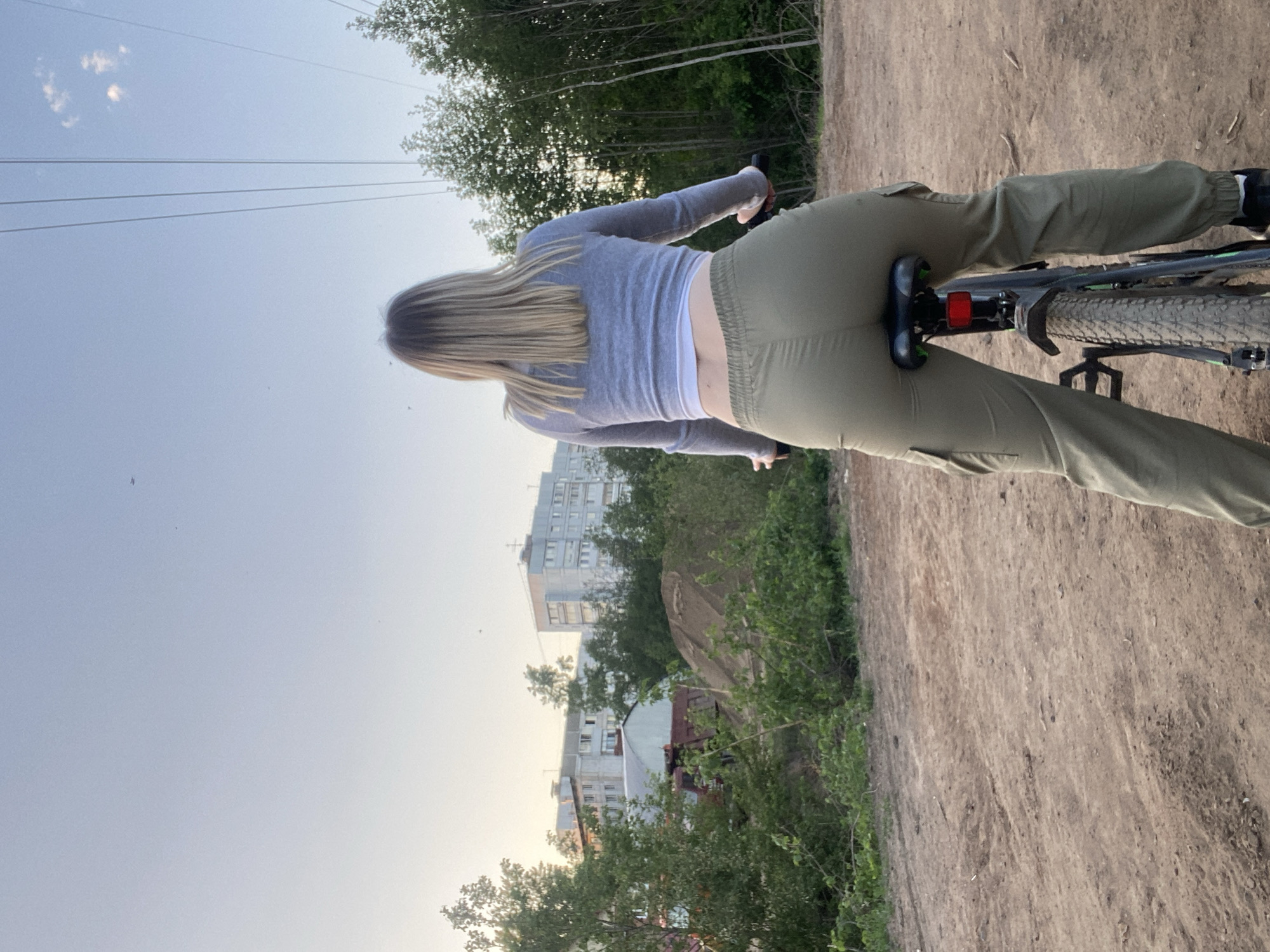 A young woman is riding a bicycle on a dirt path near a residential area in what appears to be Chaikovsky, Russia. She is wearing a gray long-sleeved shirt and olive green pants. The bicycle is a mountain bike. The background includes trees, residential buildings, and power lines. The scene appears to be taken at dusk or during sunset. The path is dirt, and the area around it is green with trees and bushes.