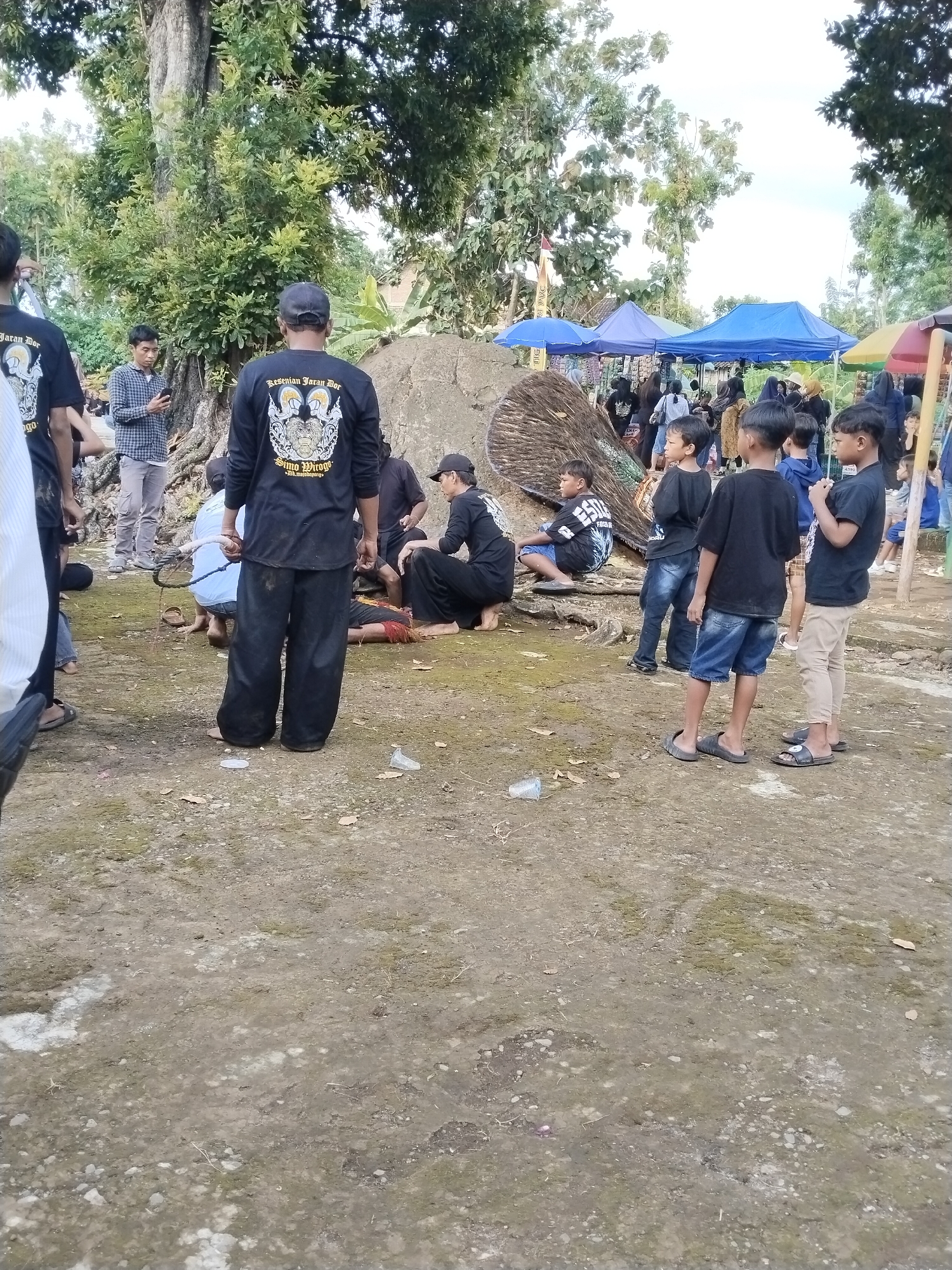 The scene is outdoors in Mojodoyong, Indonesia. Several people are gathered on a dirt and gravel surface, with trees and tents visible in the background. A group of boys stand to the right, watching an unseen event. Several men wearing black shirts with a logo on the back are standing and sitting in the center of the frame. One man is lying on the ground. A man stands with a rope in his hand.