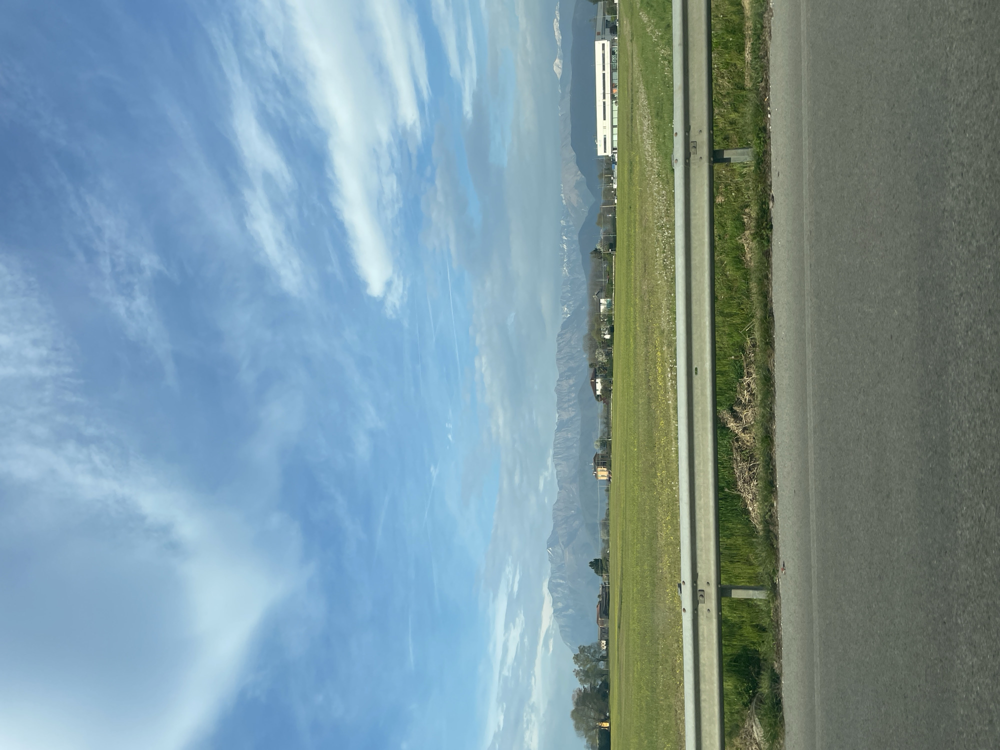 The image shows a view of a rural or semi-rural landscape, possibly taken from a moving vehicle. The foreground features a gray asphalt road and a metal guardrail. Adjacent to the guardrail is a grassy area or field, appearing green and expansive. In the distance, a mountain range is visible, with some snow on the peaks. Further off, there are some buildings, possibly houses or other structures typical of the city of Colloredo di Monte Albano / Colorêt di Montalban, Italy. The sky is blue with streaks of white clouds. The overall impression is of a sunny day and a scenic drive.