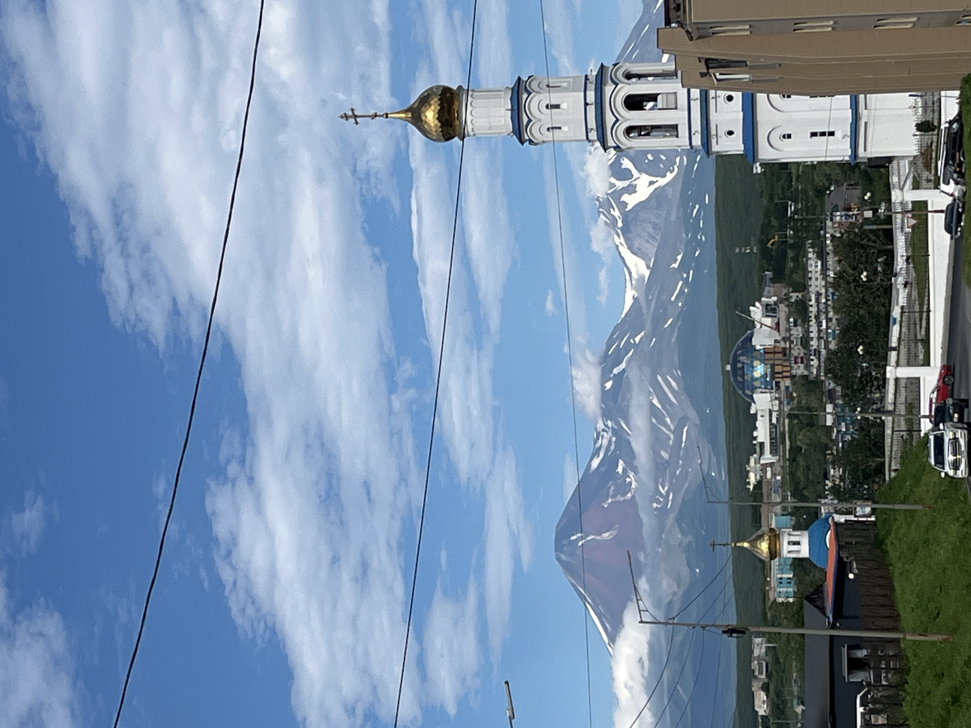 This is a photograph of a city skyline with a prominent mountain range in the background. In the foreground, a tall, white bell tower with a gold dome, topped with a cross, stands to the right. The tower has blue trim and arched openings. To the left, a smaller blue-domed church with a gold dome is visible. The city is spread out below the mountains, with various buildings, some of which appear to be residential, and a large, modern, glass-domed structure. The landscape is green and hilly. The sky is blue with scattered white clouds. Power lines are visible crisscrossing the sky in the foreground. Several cars are on the road, including a white SUV and a red car. The scene is set in Petropavlovsk-Kamchatsky, Russia, with the famous volcanoes of the Kamchatka peninsula visible behind the city.
