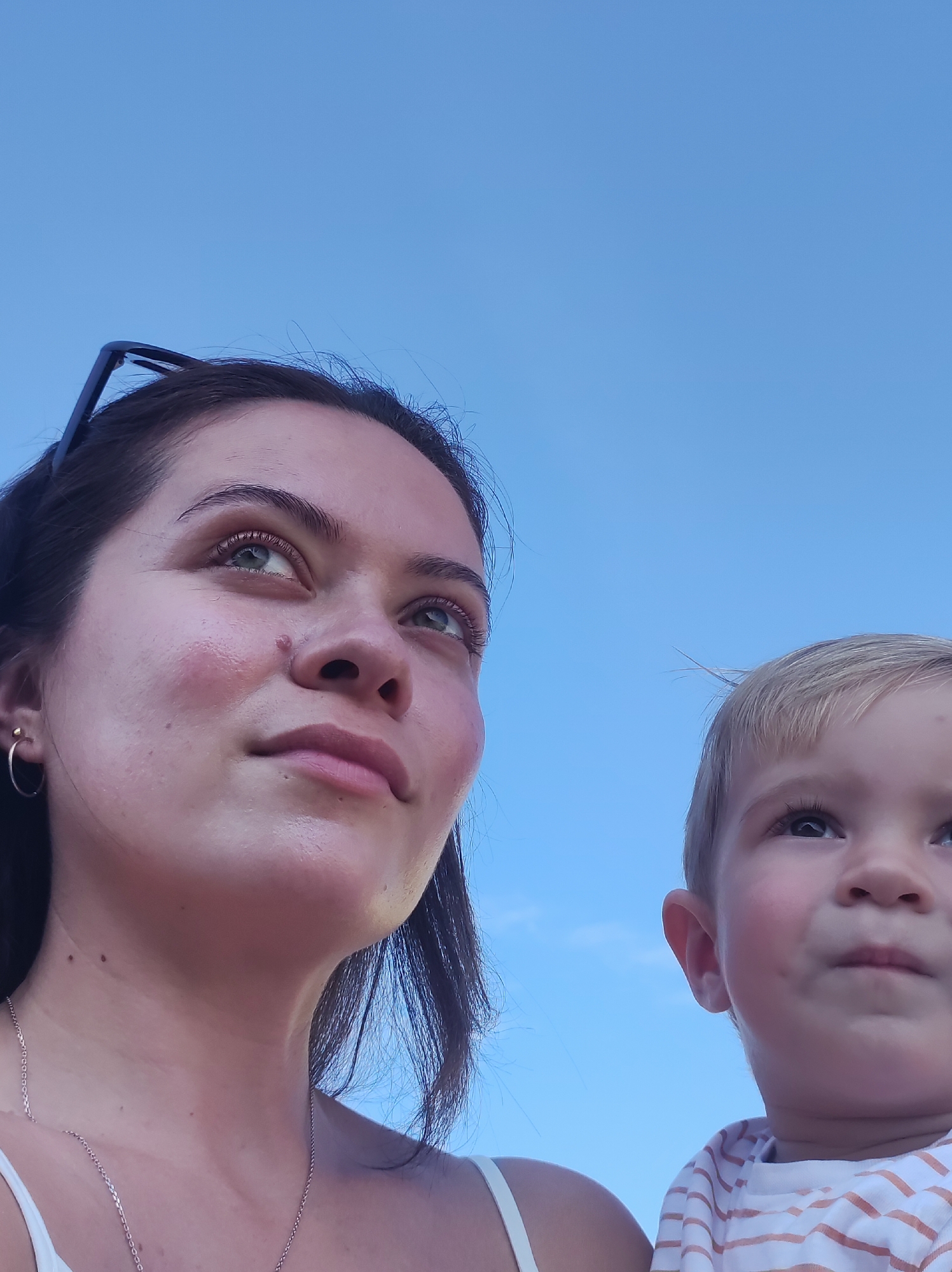 A close-up, low-angle, eye-level shot features a woman and a young child against a clear blue sky. The woman, on the left, has dark hair and fair skin. She is wearing a thin necklace and a hoop earring in her visible ear. A mole is visible on her left cheek. The child, on the right, has fair hair and appears to be looking upwards. They are wearing a horizontally striped shirt. The focus is sharp on their faces, suggesting the image was taken outdoors in daylight. There is no indication of the city of Chaikovsky, Russia, in the image.