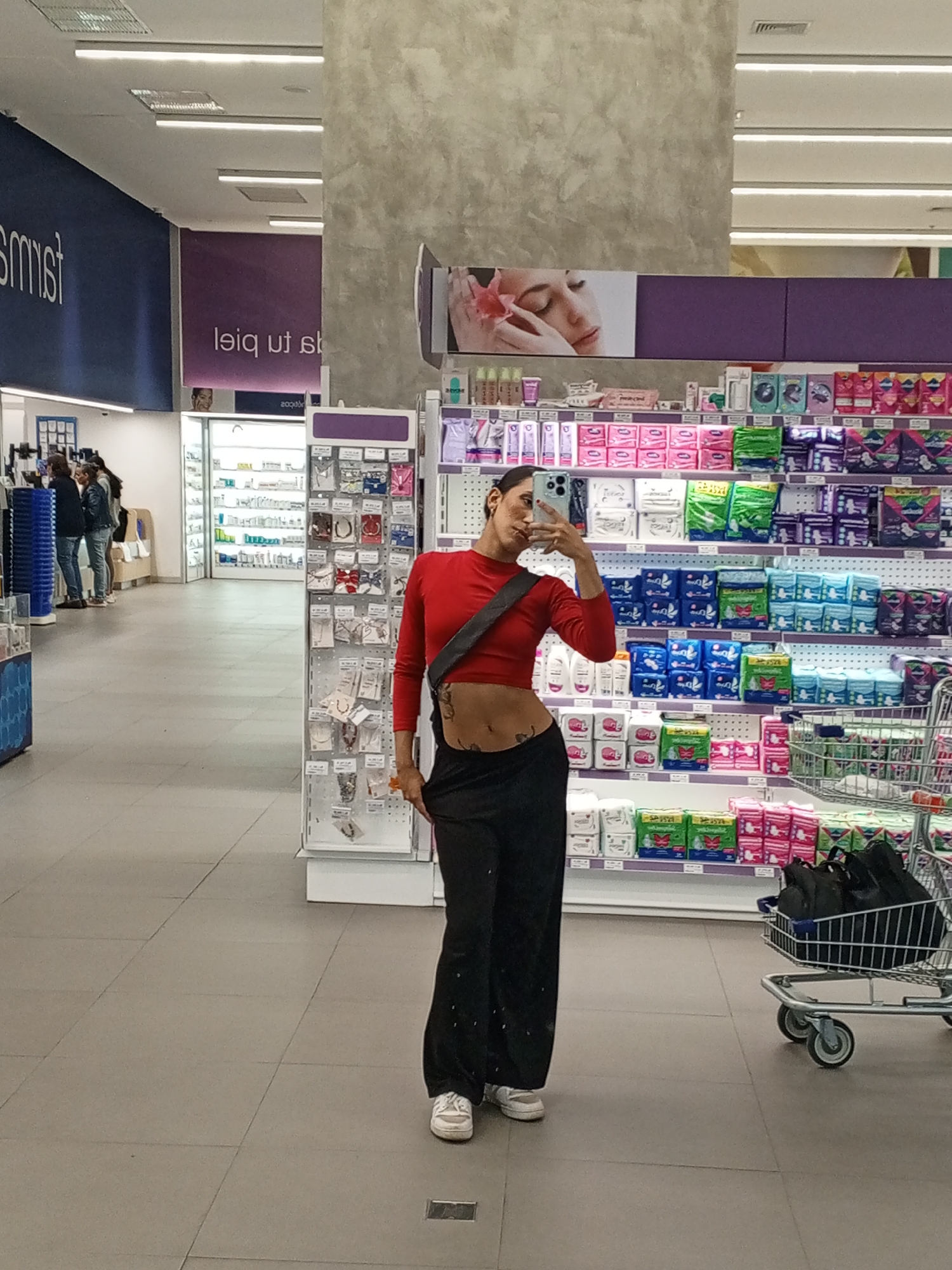 A woman with brown hair and a red crop top is taking a selfie in a pharmacy. She is wearing black pants and white sneakers. She has a black bag slung across her chest. A shopping cart filled with black bags is next to her. Behind her are shelves stocked with feminine hygiene products and other goods. Other people are in the background. The location is in the city of Porlamar, Venezuela.