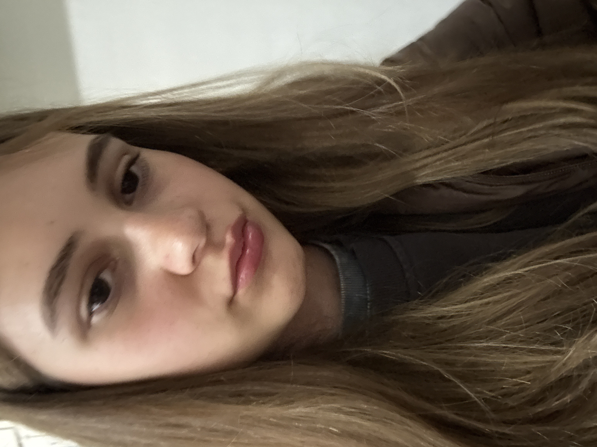This close-up portrait, taken in Panagyurishte, Bulgaria, features a young person, likely female, with fair skin and long, flowing light brown hair with visible blonde highlights. The subject's head and upper torso are captured at an angle, suggesting she is lying down or reclining.
Her face, prominent in the foreground, displays clear skin, dark, well-defined eyebrows, and dark eyes that gaze slightly upwards. She has a small nose and full lips with a natural pinkish tint. Her expression is calm and neutral.
She is wearing a dark, possibly grey or charcoal, ribbed turtleneck top, partially covered by what appears to be a dark brown or black puffer jacket. Her hair cascades around her head, filling much of the frame. The background is a plain, light-colored wall or surface. The lighting is soft and even, suggesting an indoor setting without harsh shadows. No text is visible in the image.