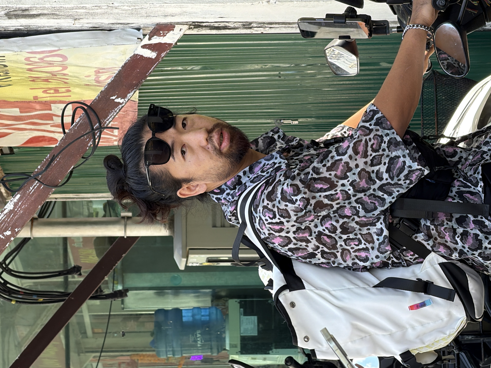 In a sun-drenched outdoor scene, a man with dark hair styled in a bun and a beard is pictured from the chest up, seated on a motorcycle. He wears black sunglasses and a short-sleeved button-down shirt with a pink and gray leopard print pattern. A white backpack with black straps is visible on his left shoulder, and a black fanny pack is worn across his chest. The man's right hand, adorned with a silver bracelet, is visible on the motorcycle's handlebar. The background features a green corrugated metal wall, a weathered wooden beam with black cables, and a partially obscured sign with red and yellow text. A blue water cooler dispenser is visible to the left. The scene appears to be on a street in Ko Samui District, Thailand, indicated by the style of the buildings and the overall tropical ambiance.