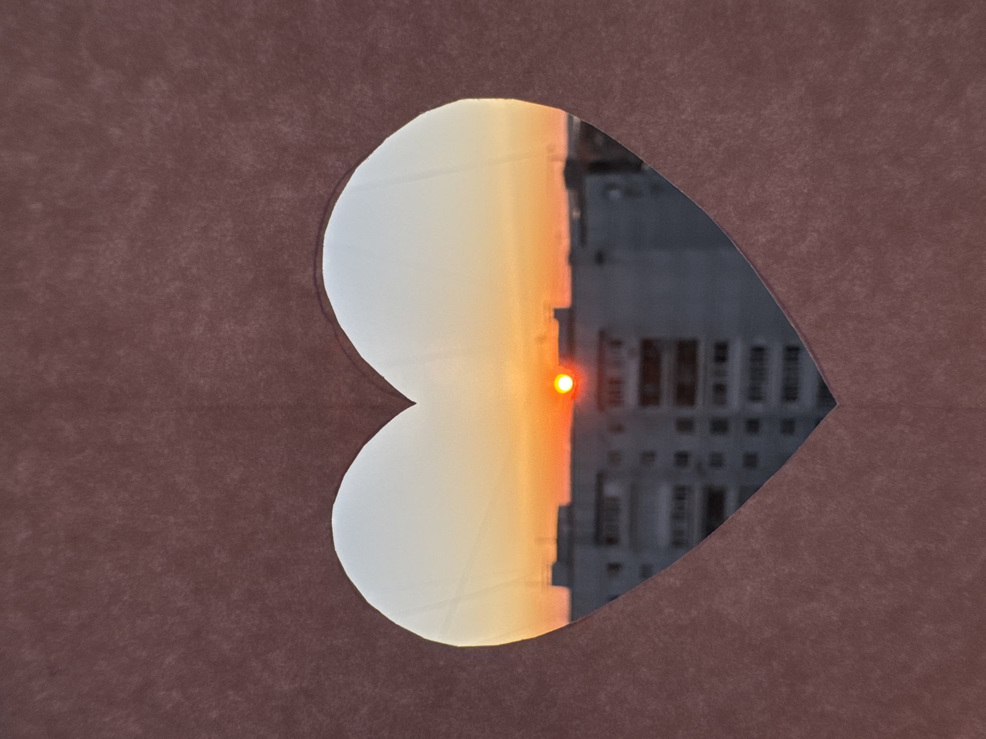 The image shows a heart-shaped opening cut into a brown material. Through the opening, a sunset is visible, with a bright orange sun low on the horizon. To the right of the sun is a gray building, its windows visible. The background is the city of Chaikovsky, Russia.