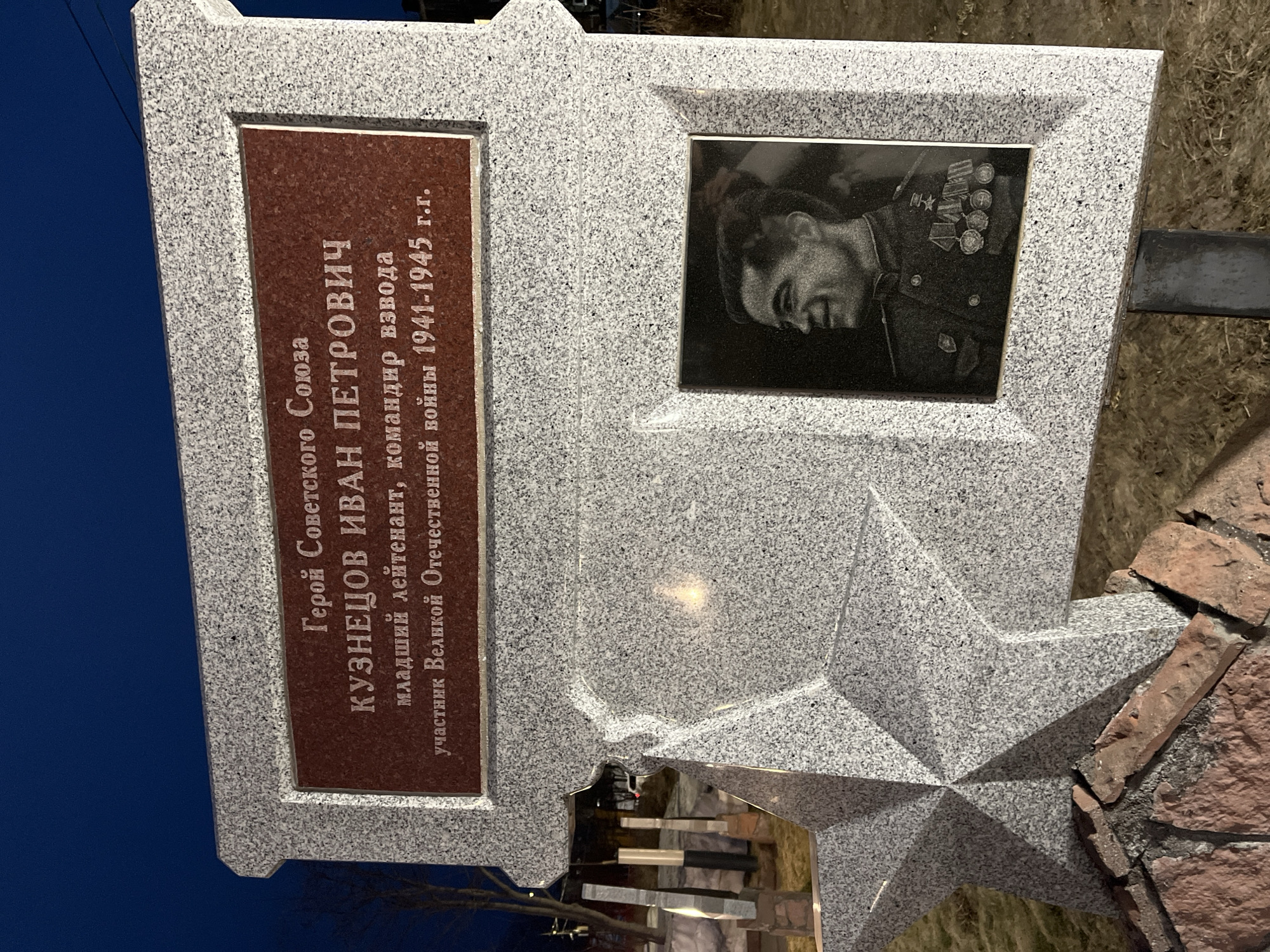 The image shows a memorial in Petropavlovsk-Kamchatsky, Russia, dedicated to Hero of the Soviet Union, Kuznetsov Ivan Petrovich. The memorial is made of light gray granite and features a five-pointed star at the bottom. A red plaque displays the inscription in Russian: "Герой Советского Союза КУЗНЕЦОВ ИВАН ПЕТРОВИЧ младший лейтенант, командир взвода участник Великой Отечественной войны 1941-1945 г.г." which translates to "Hero of the Soviet Union KUZNETSOV IVAN PETROVICH Junior Lieutenant, platoon commander Participant of the Great Patriotic War 1941-1945." To the right of the plaque is a black and white portrait of Kuznetsov in his military uniform adorned with medals. The memorial is situated outdoors with a clear blue sky in the background on the left, and on the right is some type of brick landscaping.