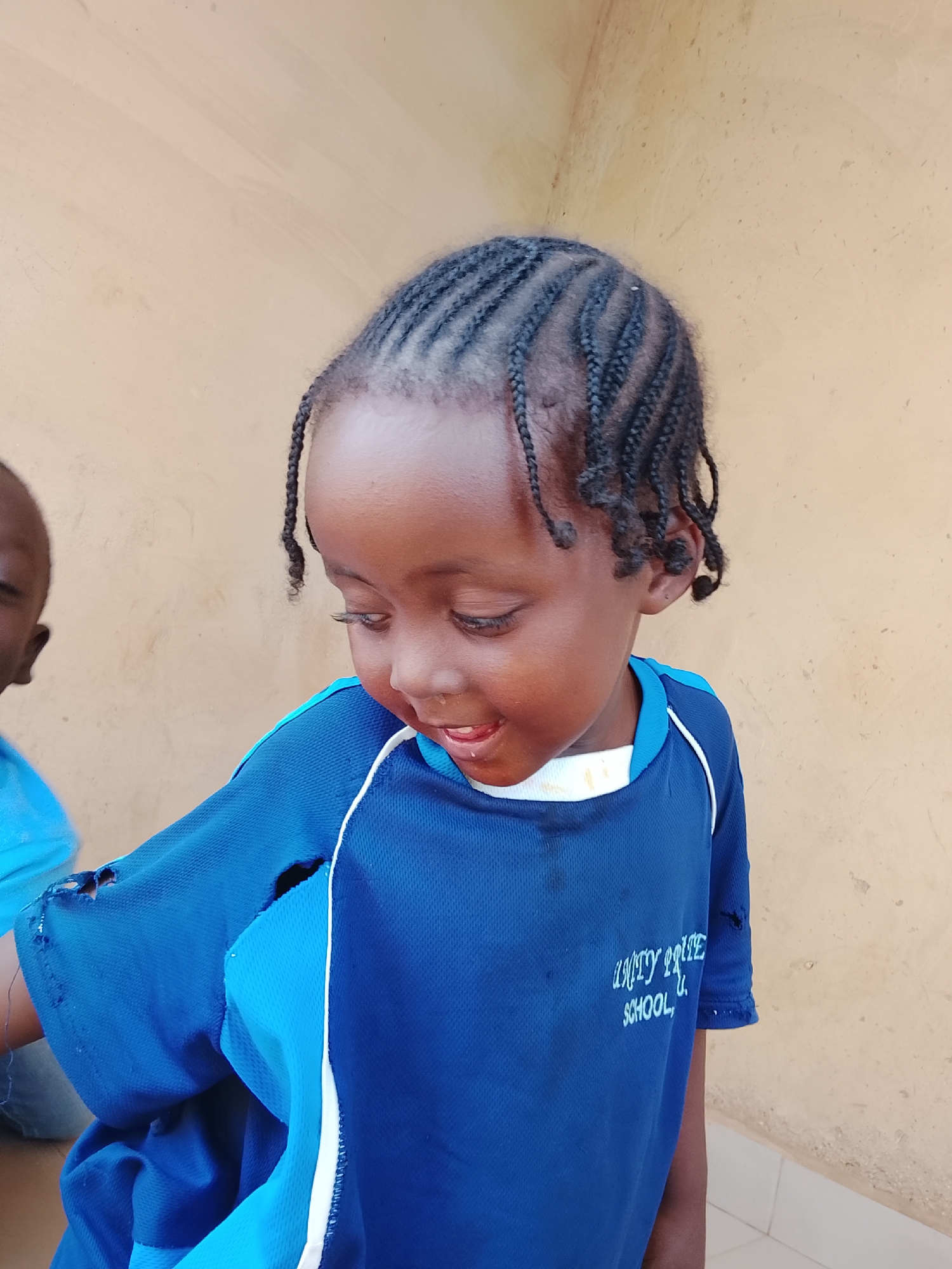 A young, dark-skinned child stands against a tan wall, looking down and to the left. The child has braided hair and is wearing a blue shirt with white trim and the words "AMITY PRIDE SCHOOL" printed on the front. The shirt has a hole near the shoulder. To the left of the child, a portion of another person wearing a blue shirt is visible. This image was taken in Kuru Dungus, Nigeria.