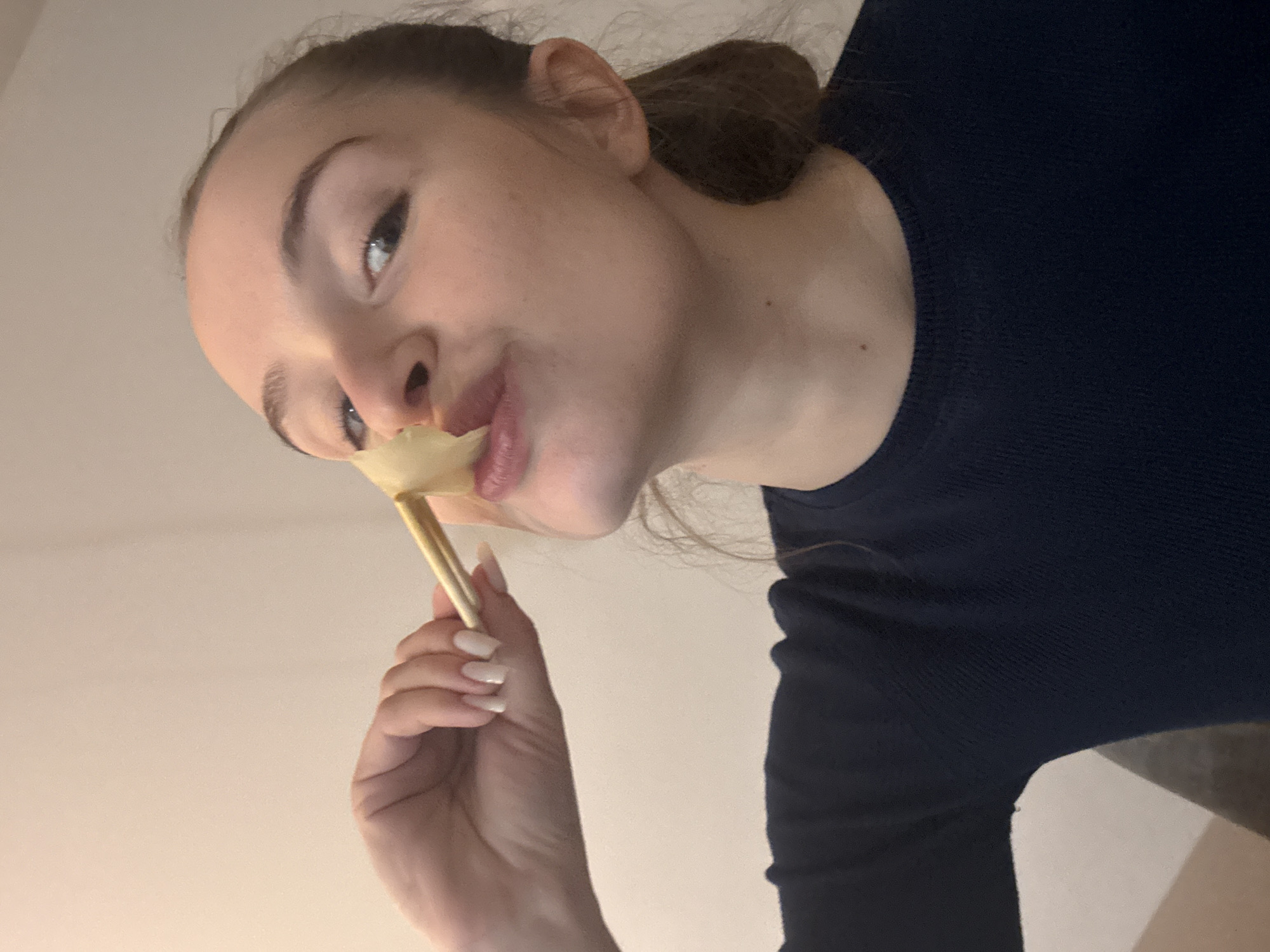 A young woman with fair skin and light brown hair pulled back in a ponytail is holding a pale yellow flower or object with a stem, close to her mouth. She is wearing a dark blue long-sleeved shirt. Her nails are manicured with a light, neutral color. The background appears to be a plain, light-colored wall. The camera angle is tilted upwards. There is no indication of location. There is no indication of city of Budva, Montenegro.