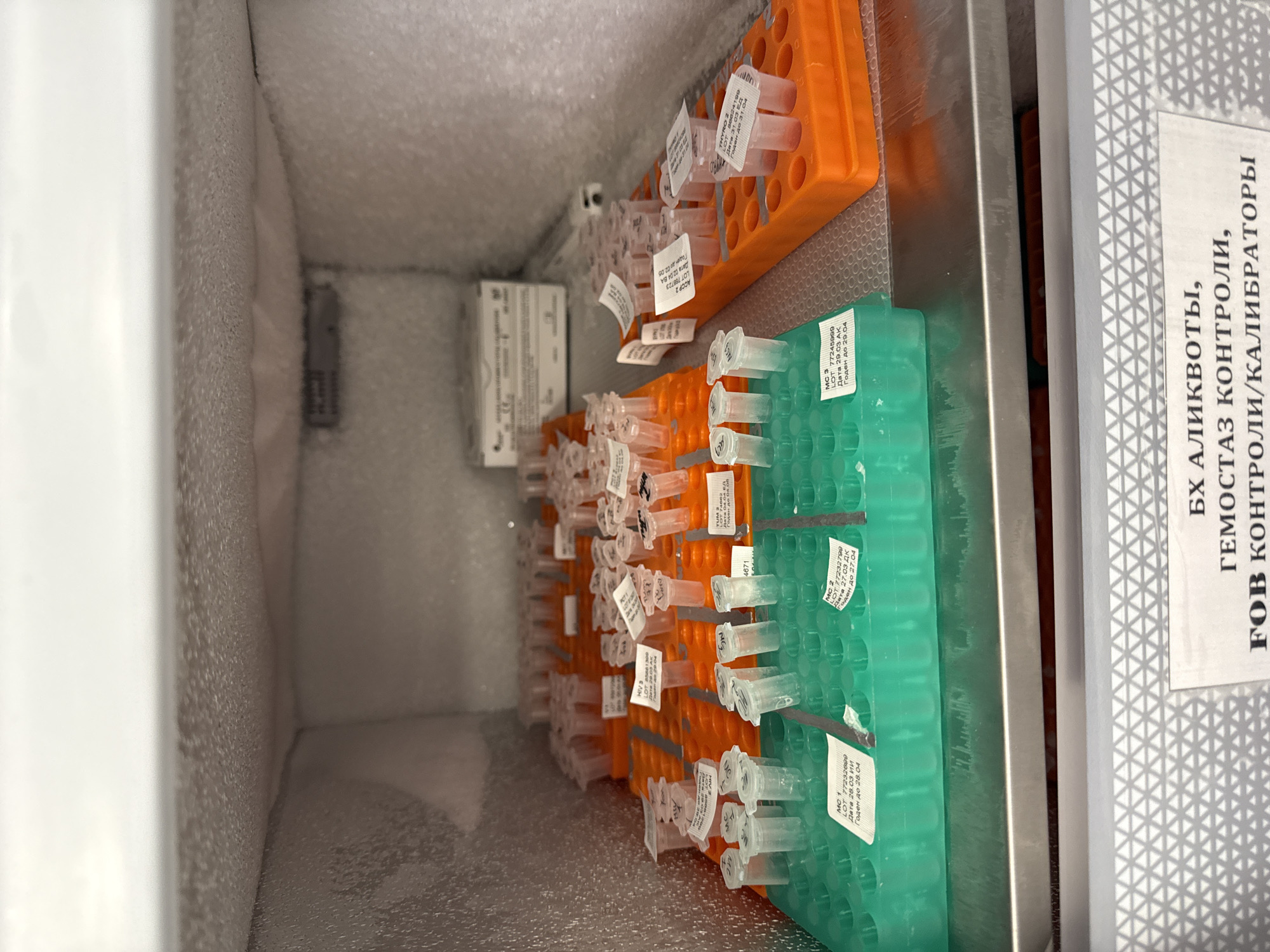The image shows a freezer compartment containing rows of small tubes held in orange and green racks. Each tube has a label affixed to it, some of which are partially visible. The labels contain text, including lot numbers and dates written in Russian. Text on a white sign visible on the right side of the image reads in Russian "Biochemical aliquots, Hemostasis controls, FOB controls/calibrators". Frost is visible on the interior surfaces of the freezer, suggesting that it is actively cooled. Based on the Russian text on the labels and the sign, the scene is most likely located in Moscow, Russia.