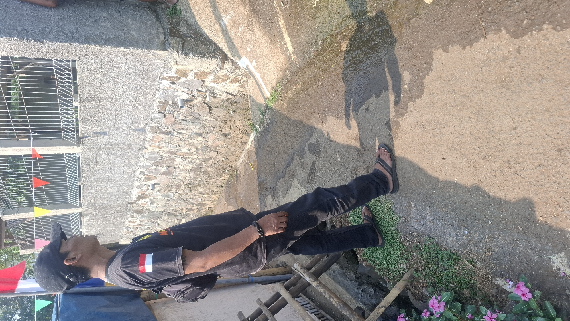 In Rahong, Indonesia, a man wearing a black t-shirt with the Indonesian flag on the left sleeve and a black baseball cap stands on a dirt path. He is looking to his right, with his body angled away from the camera. He has a black backpack slung over his left shoulder and wears black pants and dark flip-flops. To his left, a wooden structure is partially visible, and pink flowers grow along the edge of the path. Behind him, a stone and concrete wall runs along the property, topped with a metal fence decorated with colorful triangular flags. The ground appears wet in places, suggesting recent rain or water accumulation. The scene is outdoors, under bright daylight, casting distinct shadows on the ground.