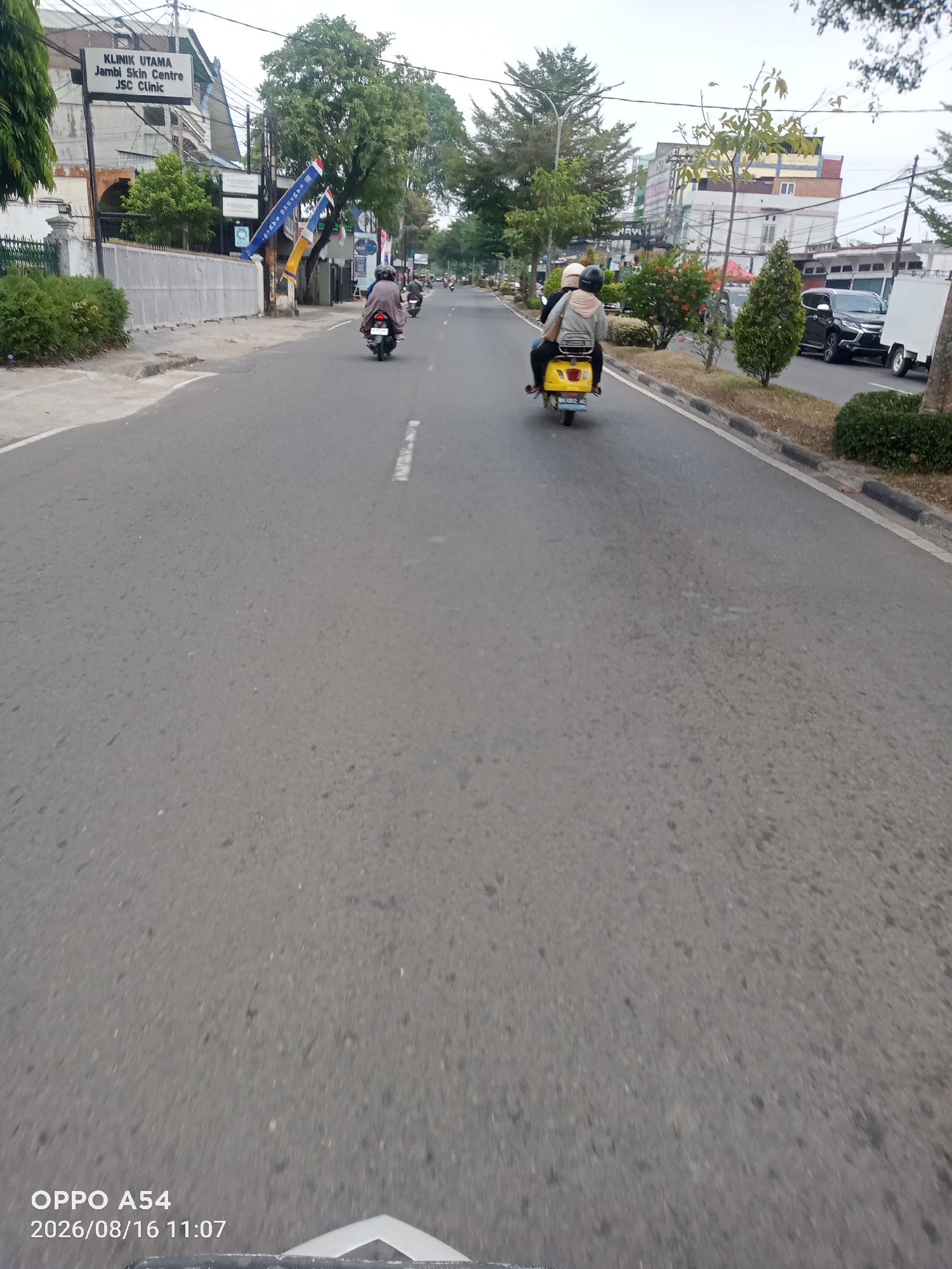 The image depicts a street scene in Jambi City, Indonesia, likely captured from a moving vehicle. The road surface is asphalt with white lane markings. On the left side of the road, a building with a sign reading "KLINIK UTAMA Jambi Skin Centre JSC Clinic" is visible, partially obscured by trees and a fence. Several motorcycles are present on the road, with two prominently featured in the foreground. One motorcycle, yellow in color, carries two individuals wearing helmets and casual attire. Another motorcycle further ahead is also occupied by a person. In the background, there are more buildings, trees, and parked vehicles, including a black car and a white truck with a trailer. The sky is overcast. The timestamp at the bottom left corner indicates "OPPO A54 2026/08/16 11:07".