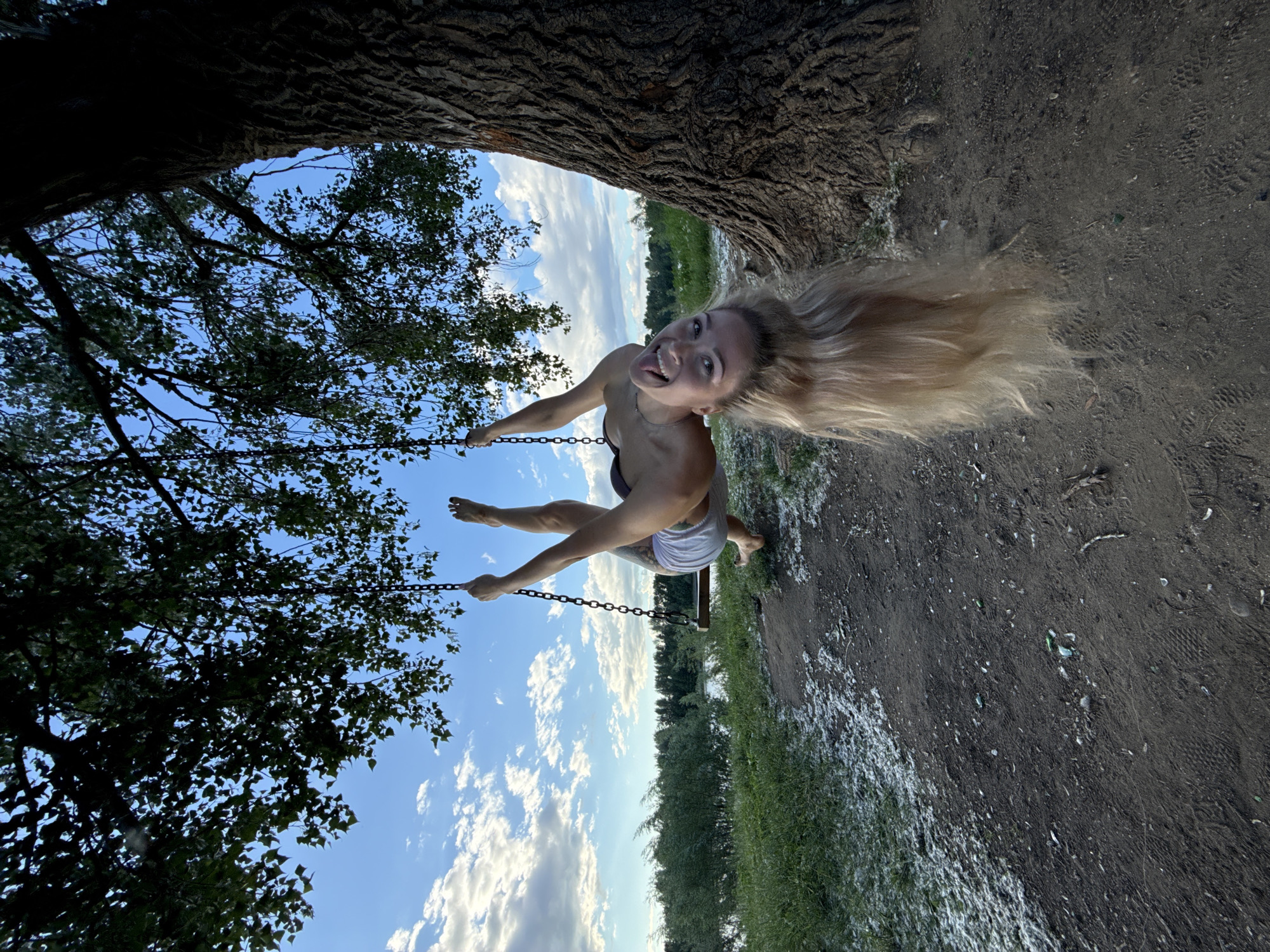 A young Caucasian woman with long blonde hair is captured mid-air on a swing, upside down, with her legs extended and her tongue playfully sticking out. She is wearing a dark-colored top and light-colored shorts. The swing is attached to a thick tree trunk, with lush green leaves of a tree partially obscuring the background on the left. The scene is set outdoors, likely in a rural or park-like environment, with a wide expanse of sky visible, dotted with fluffy white clouds. The ground beneath is a mix of dirt and grass, with some white specks that could be pollen or small blossoms. The lighting suggests it is daytime, possibly late afternoon given the angle of the sun, and the weather appears to be clear and pleasant. The woman's expression conveys a sense of joy, freedom, and lightheartedness. There is no visible text in the image. The overall impression is one of carefree fun and enjoying a simple outdoor activity.