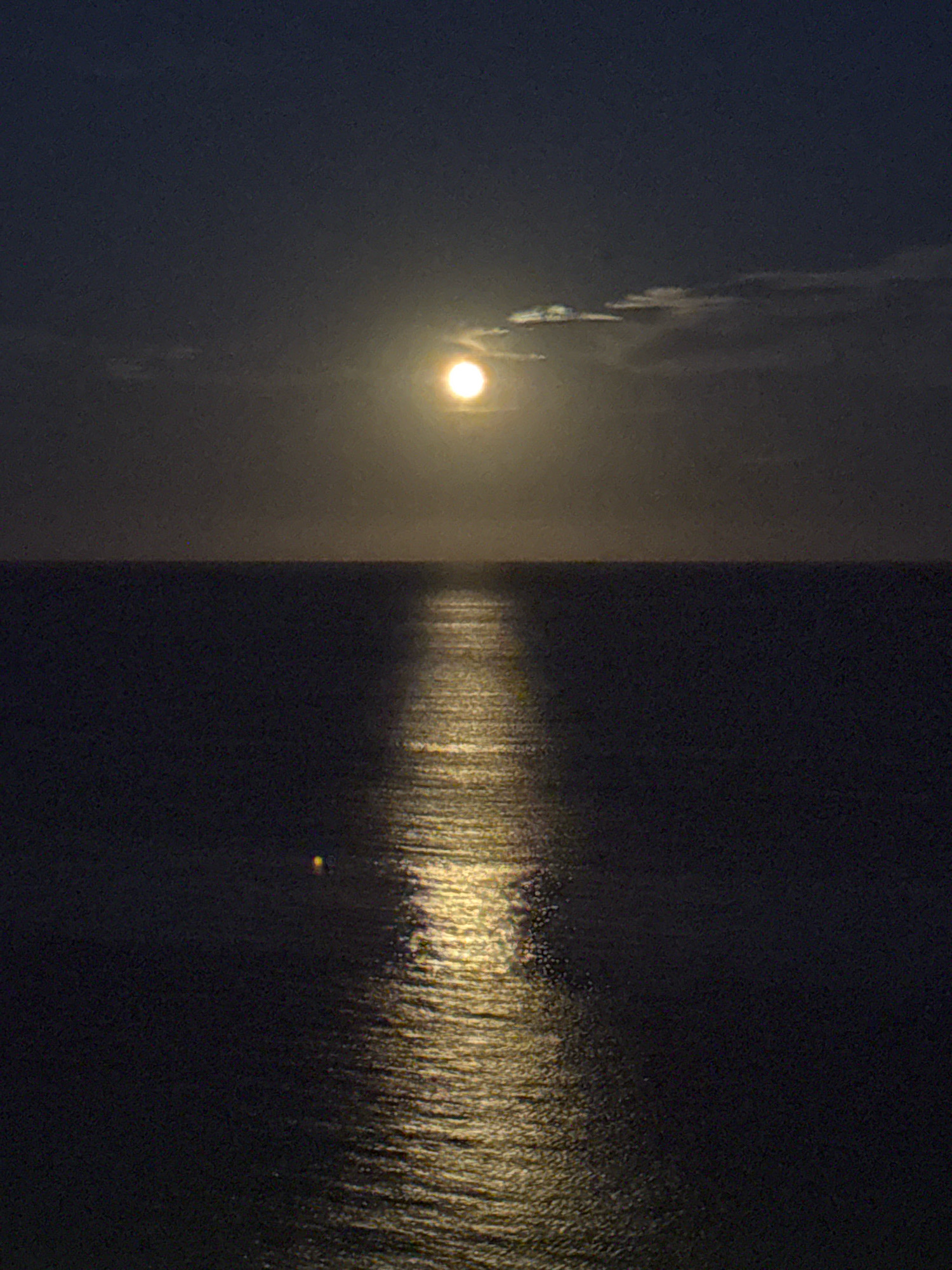 A full moon shines brightly in a dark, cloudy sky over a calm body of water. A luminous reflection of the moon stretches across the water's surface, creating a shimmering pathway towards the horizon. The scene appears to be a natural seascape, likely along the coast. Given the location provided, this image could be captured from the coastline of Calp, Spain, possibly overlooking the Mediterranean Sea with the full moon rising or setting. No people or man-made structures are visible in this photograph.