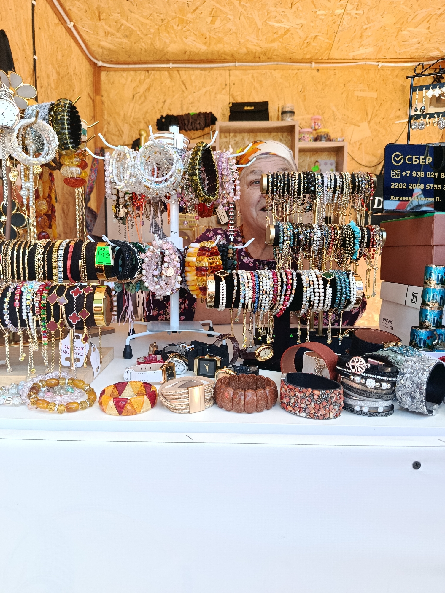 A woman with a headscarf and smiling face is standing behind a display of jewelry, which includes numerous bracelets and watches. The jewelry is arranged on stands and a table. The background appears to be a wooden wall. On the right side of the image, there is a sign with Russian text and a logo. The scene is likely an outdoor market stall in Grozny, Russia, as indicated by the presence of Cyrillic text on the signage.
