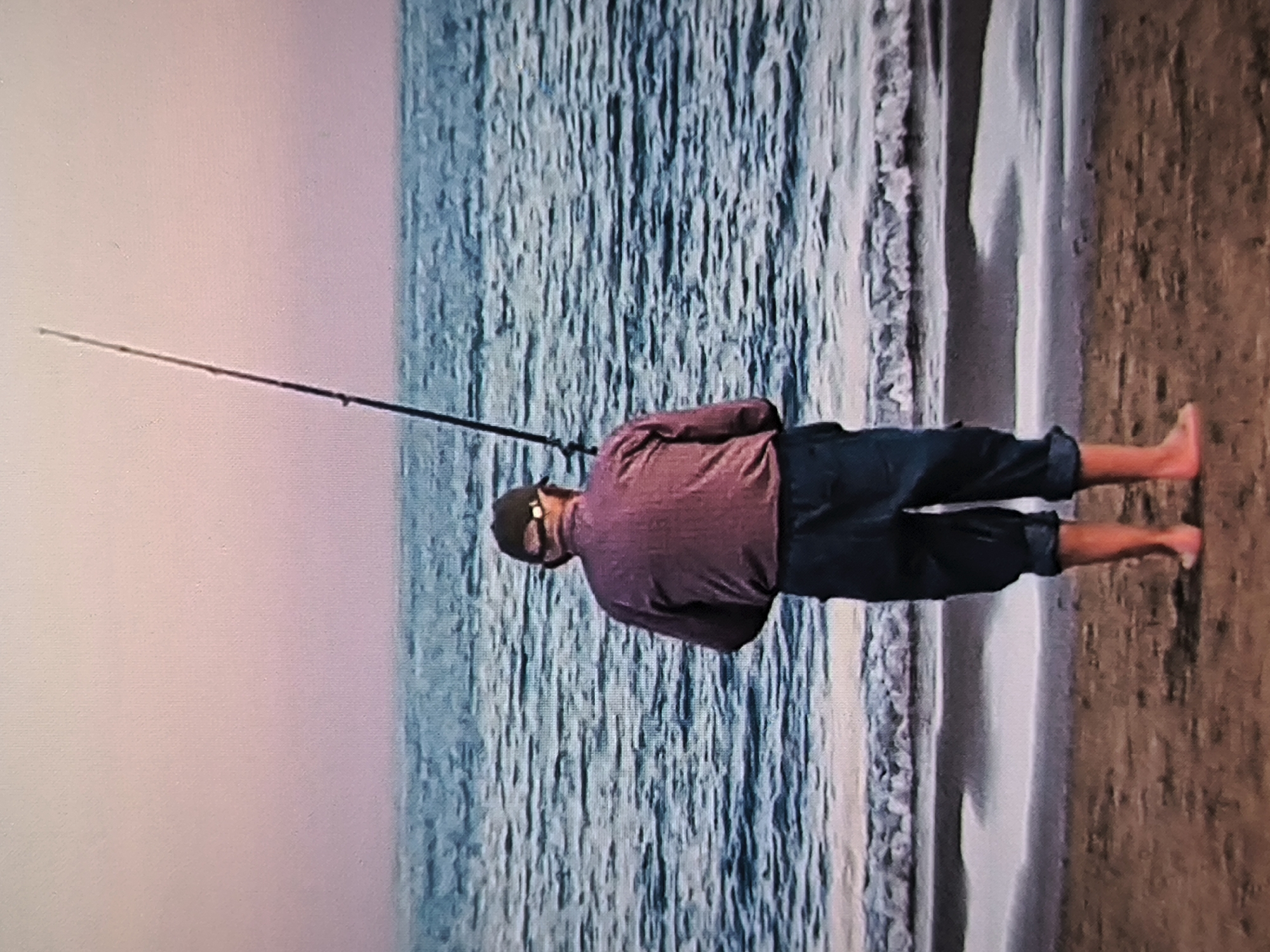 The image, when correctly oriented, depicts an adult male standing on a sandy beach at the water's edge, facing the ocean. He is viewed from behind and is holding a fishing rod that extends out over the water, indicating he is engaged in fishing.
The man is wearing a reddish-purple long-sleeved shirt, dark blue trousers or shorts rolled up to his calves, a dark baseball cap, and sunglasses. He is barefoot, standing where the gentle waves meet the shore. The ocean stretches into the distance, appearing textured with ripples and reflections. Small, foamy waves can be seen breaking softly onto the sand. The sky above presents a soft gradient of light pink and purple hues, suggesting either dawn or dusk. The scene conveys a sense of tranquility. No text is visible in the image.