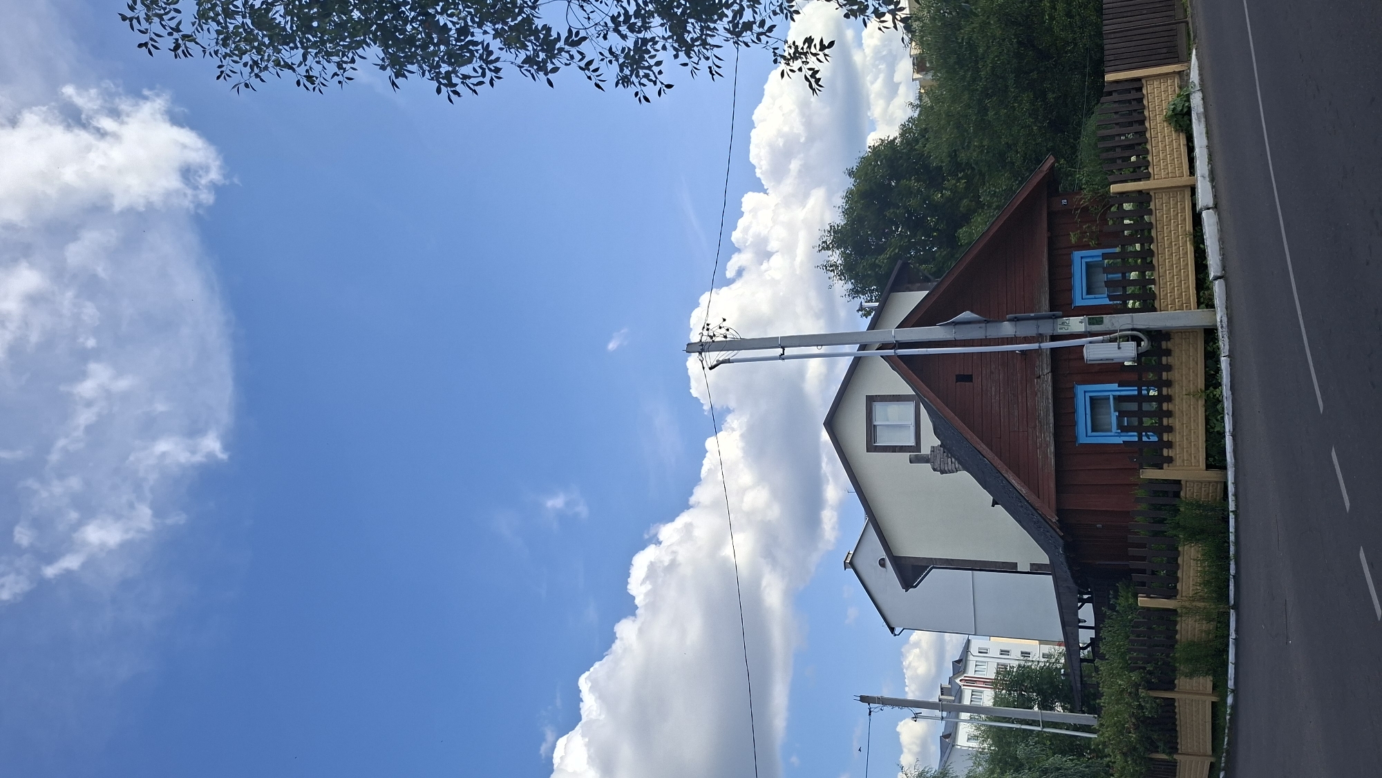 A low-angle, vertical, outdoor, daytime photograph shows a quaint wooden house with a pitched roof and blue window frames in Myadzyel, Belarus. The house is situated on a corner lot, with a two-toned fence in front of it and a paved road in the foreground. To the left of the wooden house is a larger, modern, multi-story building. The sky above is a vibrant blue with scattered white clouds and a leafy tree branch extends from the right side of the frame. A utility pole stands in front of the wooden house, adorned with electrical wires. The scene depicts a residential area with a blend of older and newer architecture.