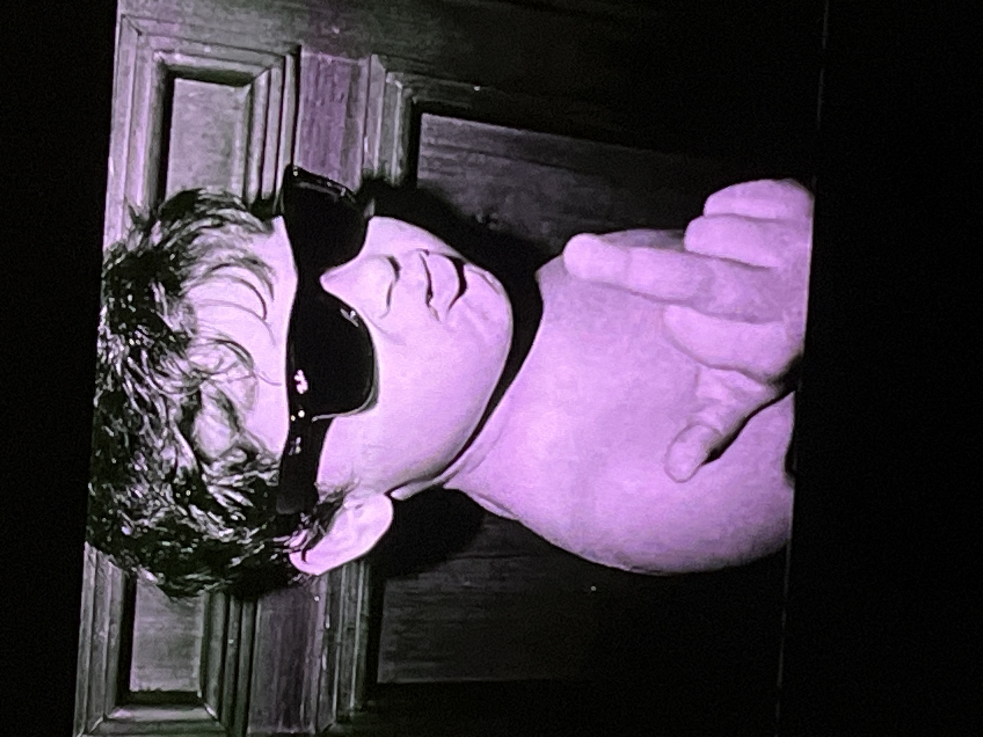 The image, rendered in a purple hue, shows a close-up of a young child wearing oversized sunglasses. The child's lips are puckered, and they are holding up a middle finger with their right hand. The background appears to be a wooden door or paneling with decorative trim. The scene is dimly lit. There is no visible indication of the child's location in Cadek, Indonesia.