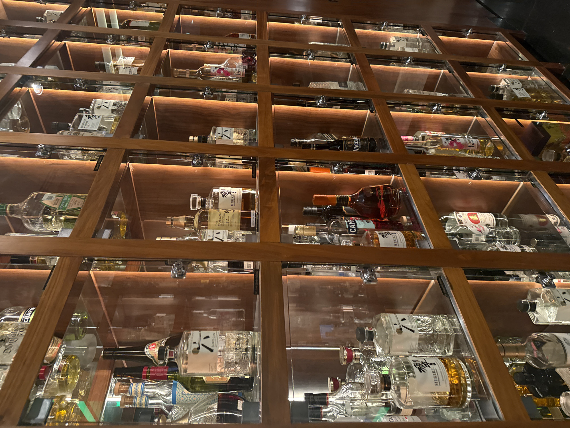 This image captures a meticulously arranged wall of illuminated wooden shelves filled with various bottles of spirits, likely within a bar, restaurant, or high-end liquor store in Puebla, Mexico. The display is structured in a grid-like pattern, with each individual compartment housing bottles behind clear glass panels and lit from within by warm, ambient lighting. The bottles are predominantly clear glass, containing a mix of golden, amber, and clear liquids, indicating a variety of whiskies, gins, and other spirits.
The scene exudes an air of sophistication and order, with no people or activity visible, focusing solely on the curated collection. Notable labels visible on the bottles, as identified by OCR, include "ROKU GIN," "HORNITOS REPOSADO," "THE YAMAZAKI 12," "HIBIKI" (appearing multiple times), "CAMP" (likely "Campari"), "SUNTORY WHISKY TOKY" (likely "Toki"), "VACCA," and "Dew" (likely part of "Plum Dew"). These labels point to a diverse selection featuring Japanese whiskies, tequila, gin, and other alcoholic beverages. The internal lighting highlights the bottles and their contents, enhancing the visual appeal of the collection. The setting suggests an evening or indoor ambiance where such a display would serve as a focal point.