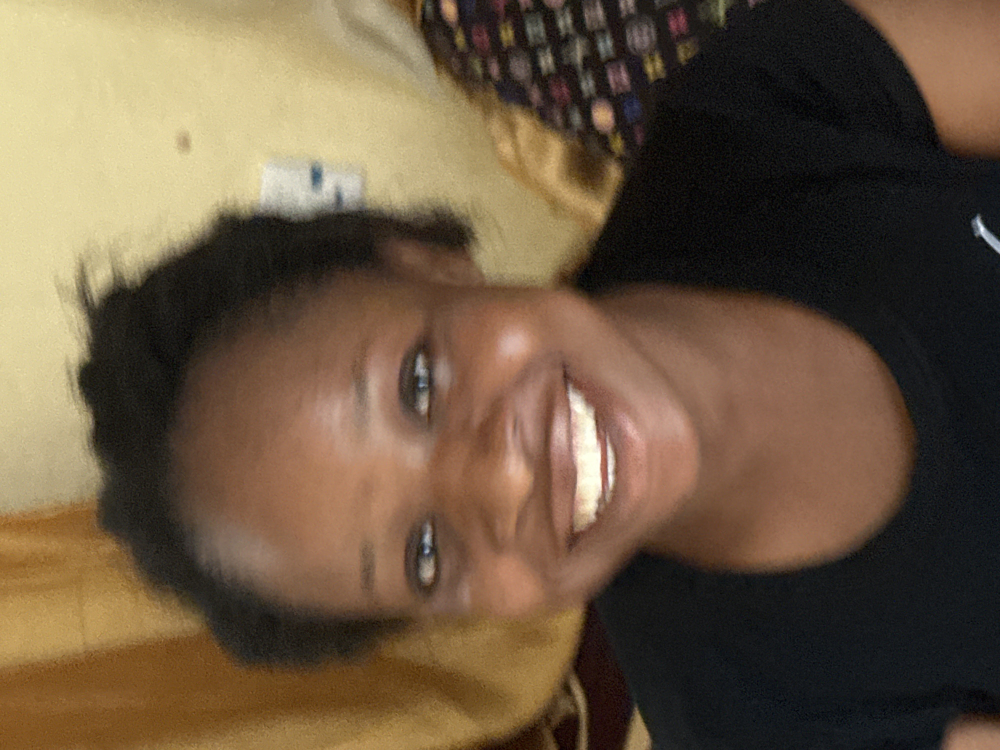 A slightly blurry portrait photograph, oriented horizontally, depicts a young woman smiling broadly at the camera. She has dark skin and short, dark, natural hair styled upwards. Her dark eyes are open and focused on the viewer, and her wide smile reveals her teeth. She is wearing a plain black t-shirt or top.
The background is an indoor setting featuring a light-yellow or beige wall. To the left of her head, some blurry details on the wall might be an electrical outlet or switch. Behind her upper shoulder and head, a dark fabric with a multi-colored, abstract pattern is visible, possibly a blanket or part of a cushion. The overall lighting appears soft and natural, illuminating her face evenly. Her expression conveys happiness and cheerfulness. The image captures a moment of joy, likely taken in Ikot Ekpene, Nigeria. There is no visible text.
