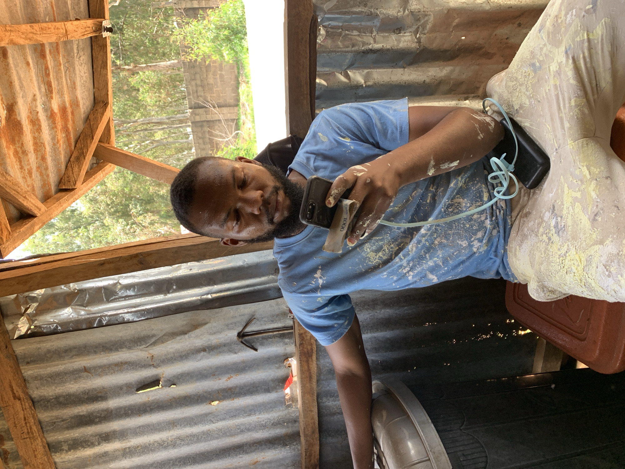 A man of African descent with dark skin, a beard, and short black hair is seated in an open-air shelter. He is wearing a light blue t-shirt with white paint splatters and light-colored pants also splattered with yellow paint. He is holding a smartphone in his right hand and a wad of cash, which appears to be Nigerian Naira, in his left hand, near the phone. A black portable charger with a light blue cord is resting on his lap. The shelter has a corrugated metal roof and walls, with wooden beams supporting the structure. Behind the man, there is foliage and a brick wall, suggesting an outdoor environment, likely in Bukuru, Nigeria. The scene appears to be in a rustic setting, possibly a workshop or a casual outdoor gathering space.