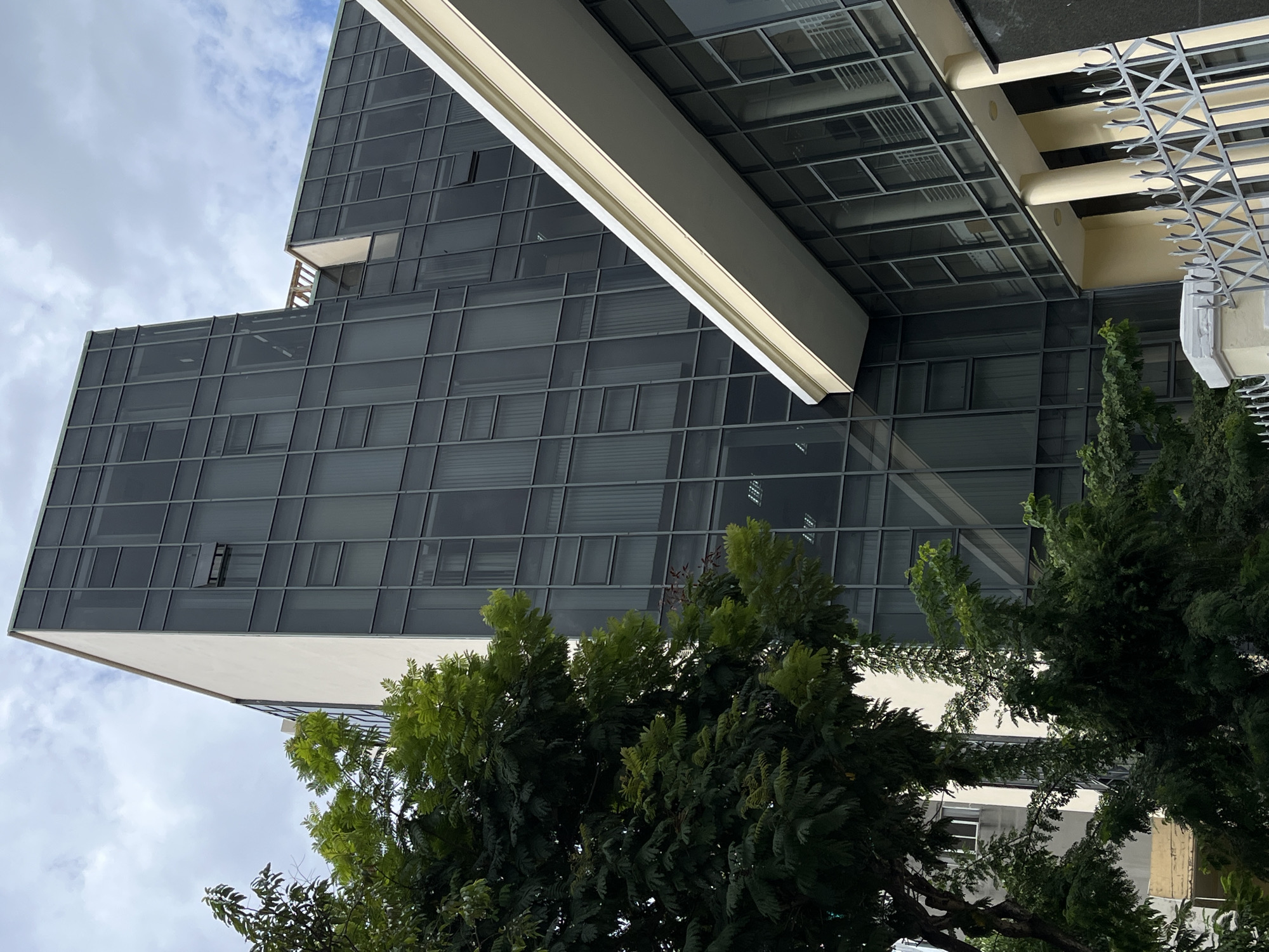 A low-angle, rotated photograph showcases a modern, multi-story building with a striking dark-tinted glass facade, partially veiled by vibrant green trees, set against an overcast sky. The architectural structure, located in Da Nang, Vietnam, features a grid pattern across its glass panels and is accented by light-colored frames. The building exhibits an angular, possibly stepped design, with sections jutting out, and at least two windows are noticeably ajar.
A long, light-colored beam or overhang extends diagonally across the upper right of the frame, adding to the geometric lines of the composition. To the far right, another part of a structure displays cream-colored pillars and a decorative metal railing with pointed elements. Lush, dense green trees, consistent with a tropical environment, occupy the foreground and midground, their foliage varying in shade and obscuring parts of the building. The sky, visible in the upper left, is filled with soft, diffused light from scattered clouds, indicating daytime. The scene is static, with no human activity or visible text.