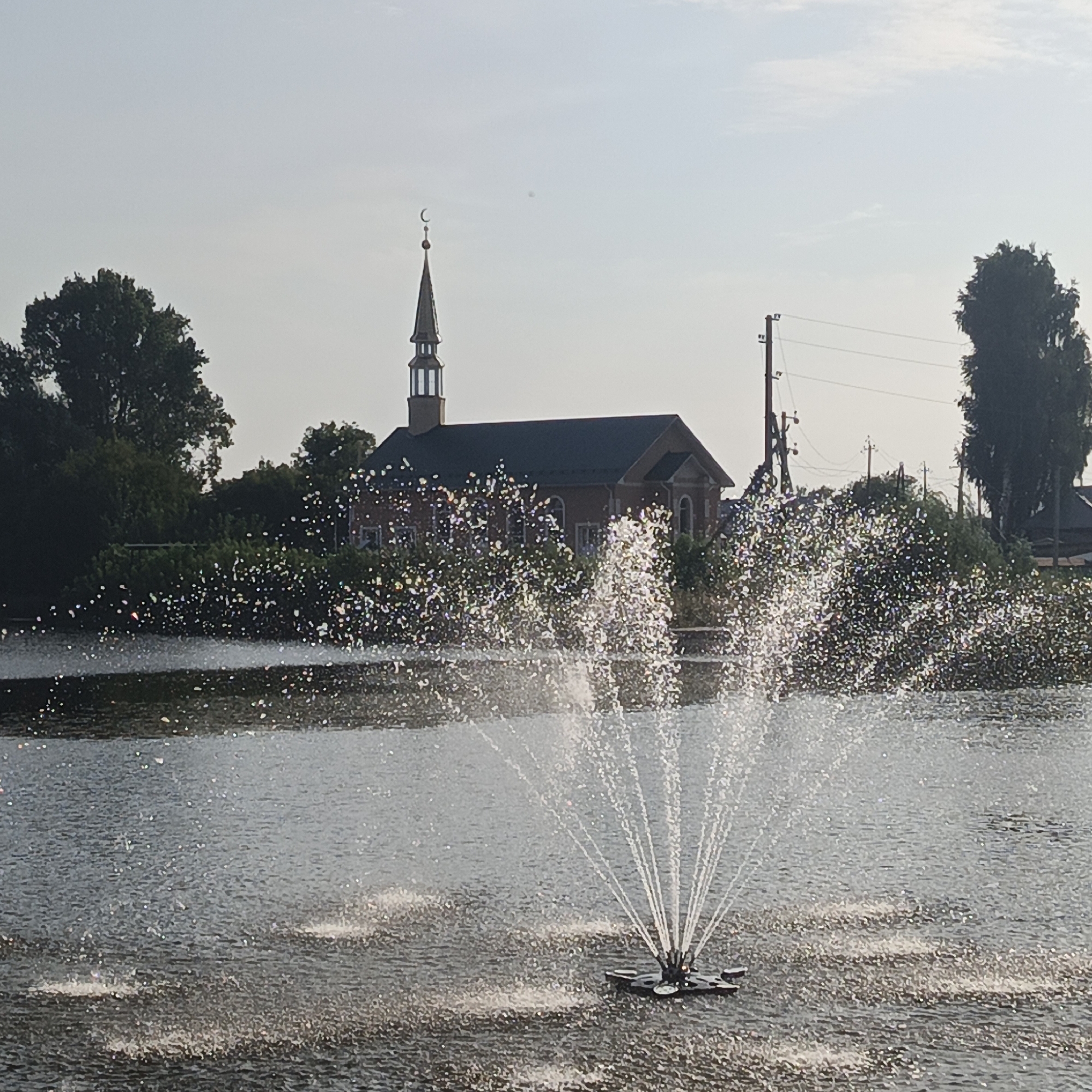 The image depicts a body of water with a fountain spraying upwards in the foreground. In the background, a brick building with a dark roof and a tall, narrow steeple topped with a crescent moon and star is visible. This building is identified as a mosque and is located in the city of Kambarka, Russia. Trees and utility poles are scattered throughout the background, and the sky is overcast. The water surface is rippled, and the fountain spray is catching the light, creating a shimmering effect.
