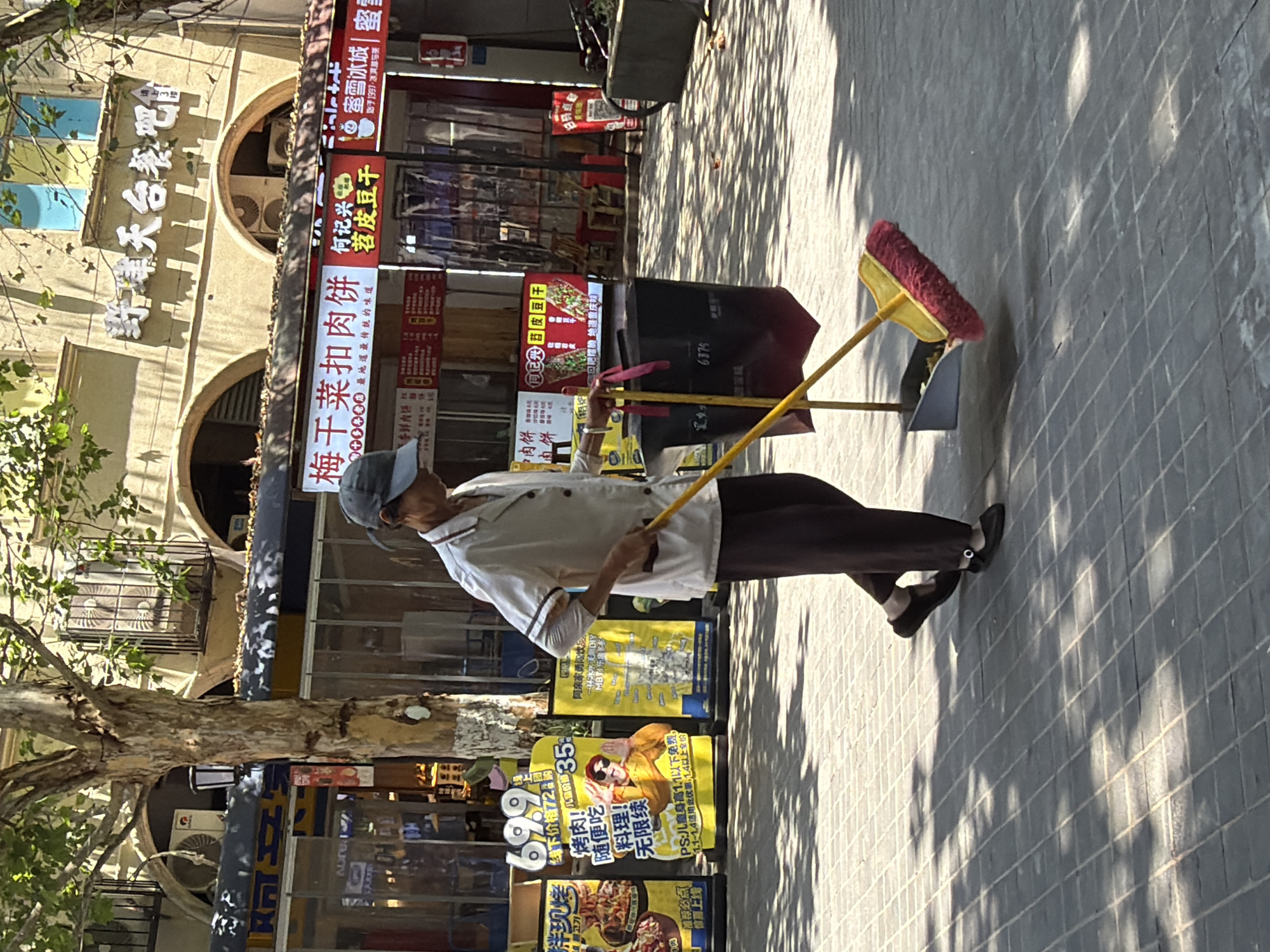 An elderly East Asian individual, wearing a white collared shirt with dark trim, dark pants, and a grey baseball cap, is sweeping the pavement with a yellow-handled broom with a pink brush. The individual is walking from left to right across the frame. To the right of the sweeper is a large black trash bag attached to a metal pole. In front of the trash bag is a dustpan containing debris. The scene is outdoors on a sunny day, with dappled sunlight and shadows from trees falling on the tiled pavement. Behind the sweeper, a building with arched windows and signage is visible. The signage includes Chinese characters, with one prominent sign featuring images of food and the text "梅干菜扣肉饼" and another sign with "69.9" and an image of a person wearing sunglasses. The general setting appears to be a commercial area or street in the city of Chongqing High-tech Zone, China.