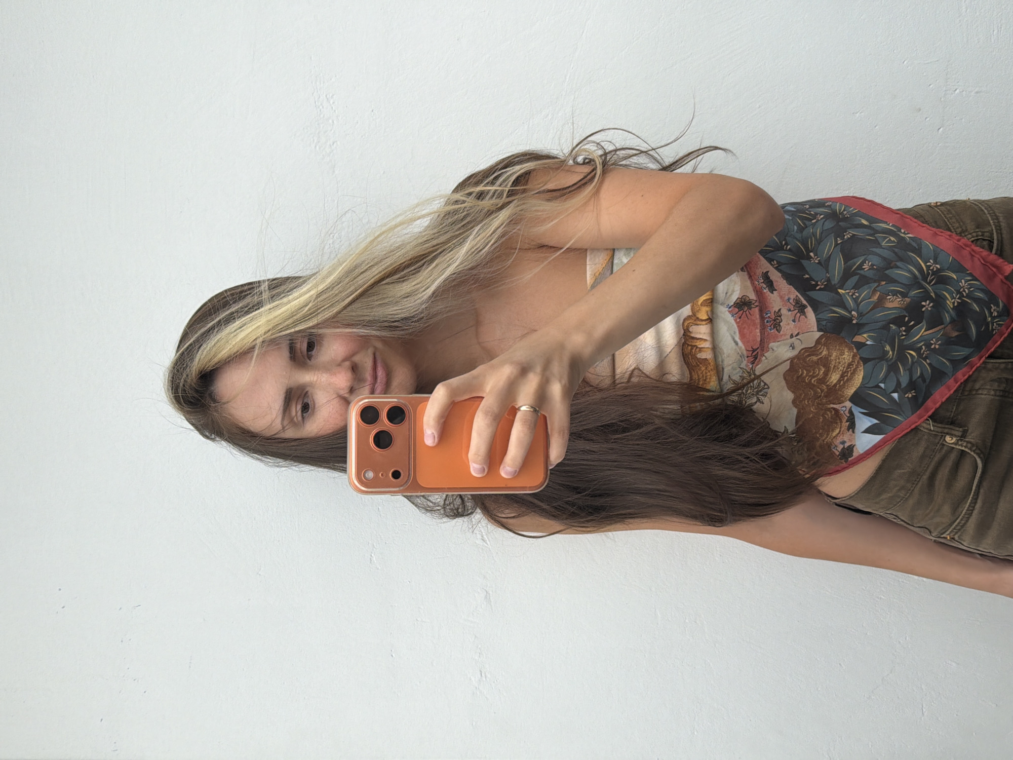 In Khimki, Russia, a young woman with long, wavy brown and blonde hair stands against a plain white textured wall. She is holding an orange-cased smartphone in her right hand, taking a selfie. Her left arm is bent, with her hand resting on her hip. She is wearing a tube top with a patterned design and brown shorts. A thin gold ring is visible on her right ring finger. The lighting is bright and even, suggesting an outdoor setting or a well-lit interior.