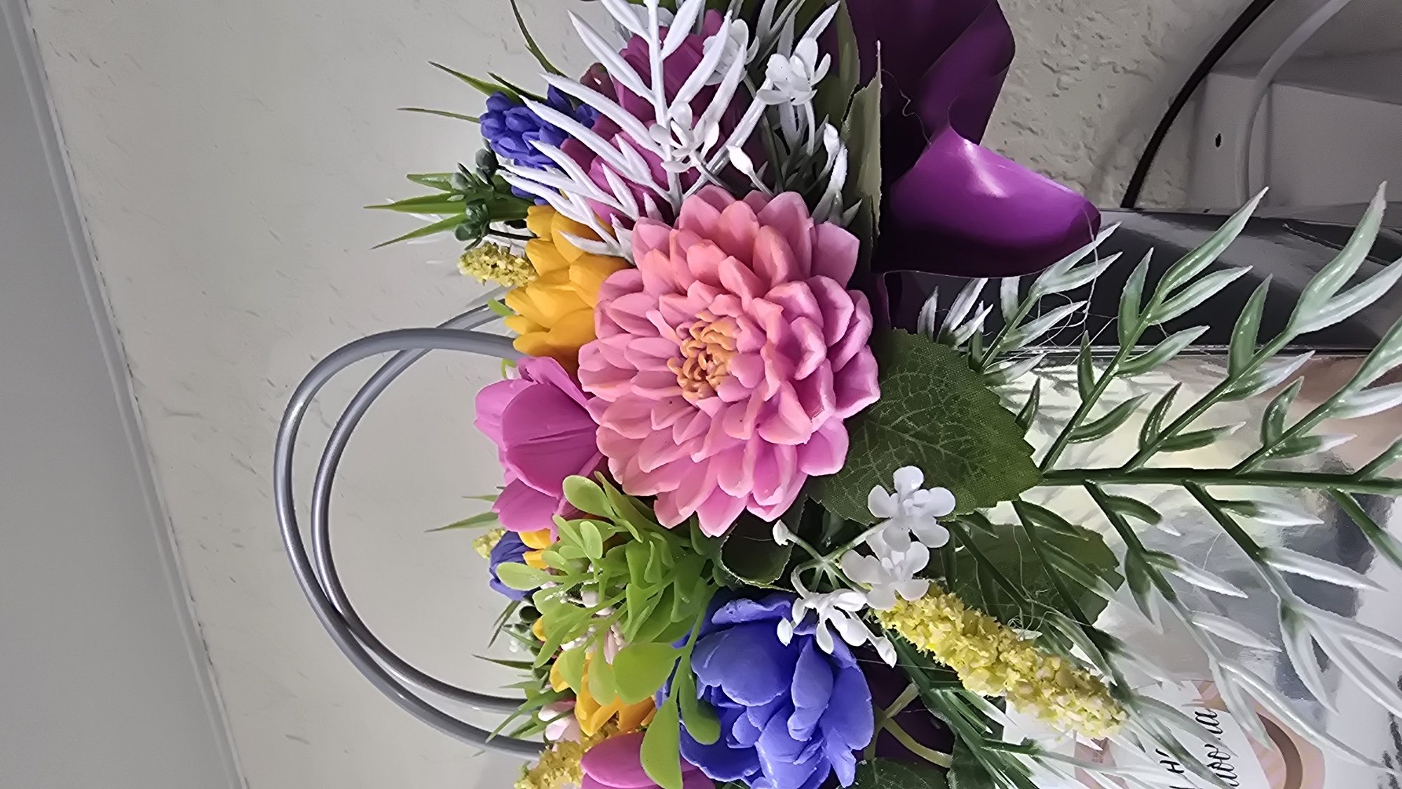 This image presents a close-up view of a vibrant bouquet composed of artificial flowers, likely crafted from soap or a similar polymer. The arrangement is held within a reflective, silvery metallic container that features a prominent, stylized handle formed by two overlapping grey circular arcs.
The bouquet showcases a variety of blooms: a large, central pink dahlia-like flower with a yellow-orange pistil, several smaller purple/blue peony-like flowers, bright yellow tulip-like flowers, and hot pink tulip-like flowers. Interspersed among the flowers is a diverse selection of artificial foliage, including small green succulent-like clusters, thin spiky green leaves, white and light green variegated foliage, small white five-petal blossoms, and clusters of tiny yellow florets. A dark purple ribbon or fabric element is visible wrapped around the right side of the container. On the lower left portion of the container, some light-colored text, partially obscured and appearing to be in Cyrillic script (e.g., "Навооа" or "Навона"), is discernible.
The background consists of a plain, light-colored wall with a textured finish, possibly stucco, against which the bouquet is set. A horizontal line, indicating a ceiling or molding, is visible across the upper portion of the wall. In the lower right background, faint outlines of an electrical cable or device suggest an indoor environment. This scene is located in Kazan, Russia.