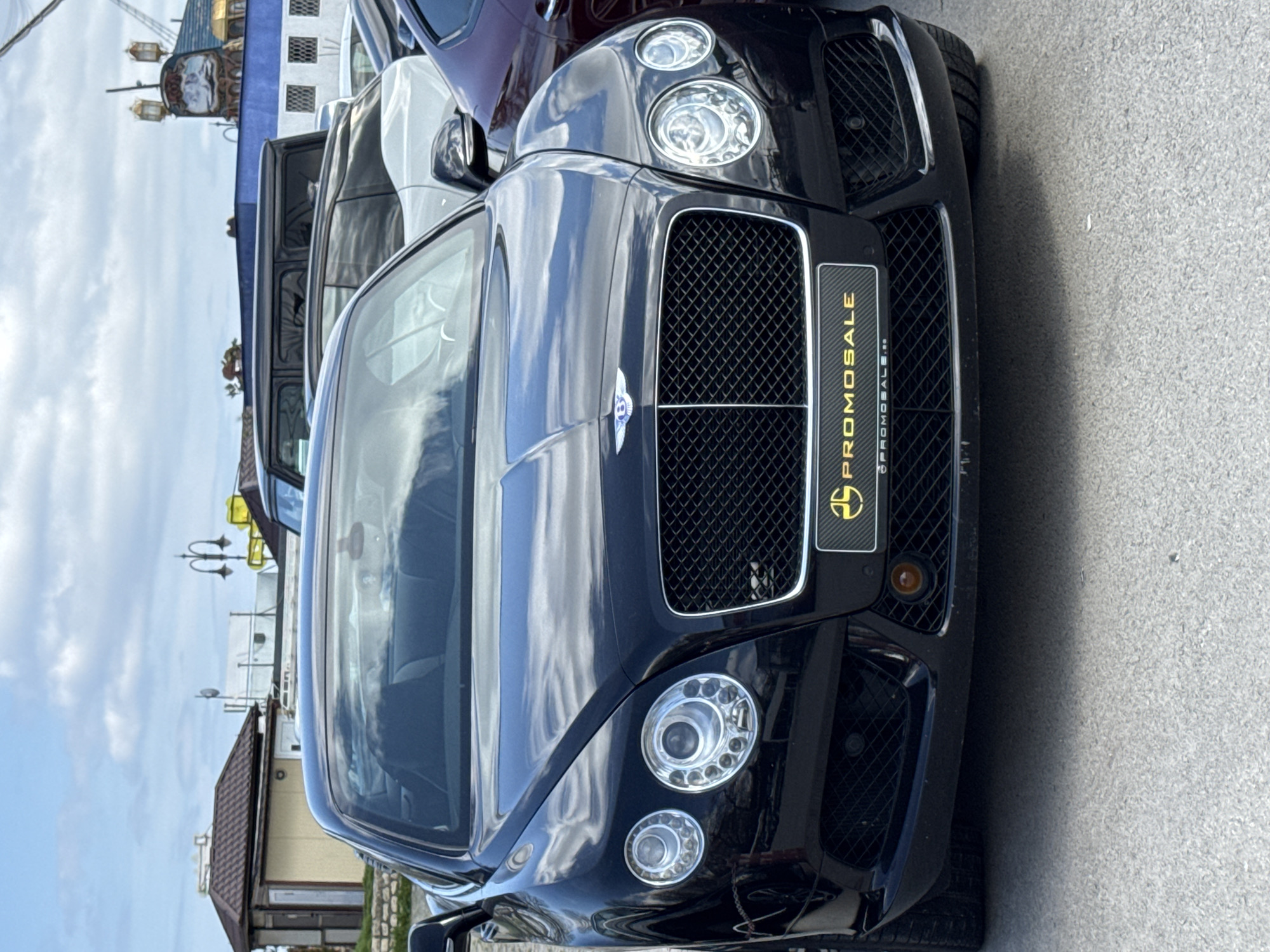 The image shows the front of a black Bentley automobile. It features the car's iconic grille with the Bentley emblem, distinctive headlights, and a license plate holder with the word "PROMOSALE" vertically. Other vehicles are visible in the background, though less distinctly. The setting is likely a parking area or street. I am unable to identify the specific location as Varna, Bulgaria without further contextual clues within the image.