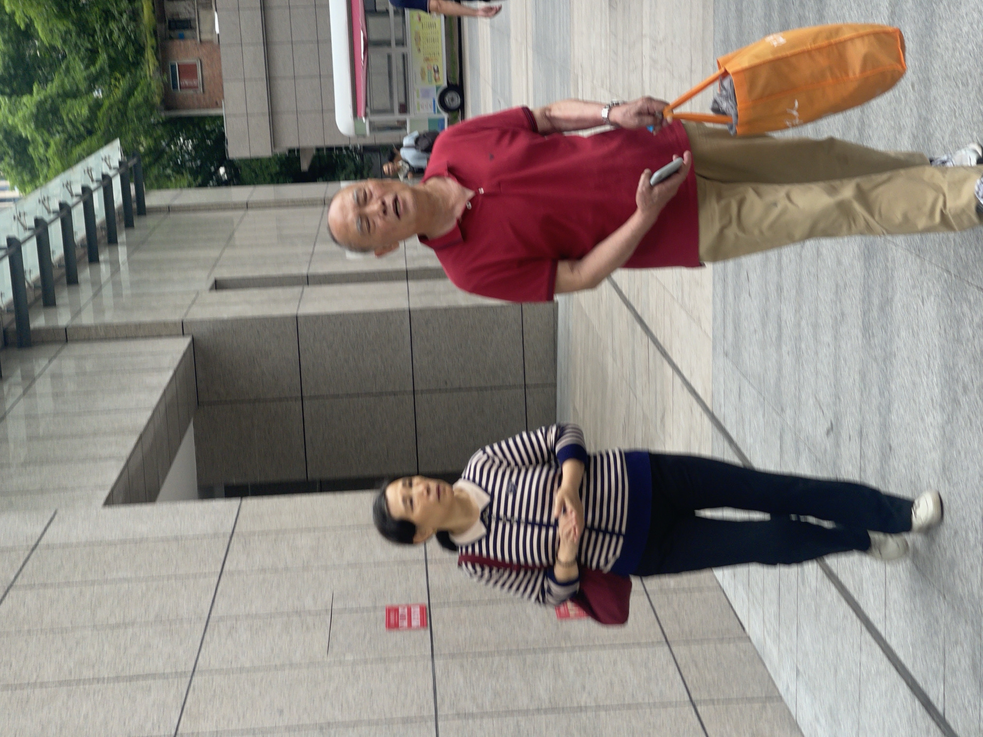An elderly man in a red polo shirt and khaki pants walks with an orange tote bag and a cell phone in his hands. Nearby, an elderly woman wearing a striped shirt and black pants stands with her hands clasped. The scene appears to be in the Yuzhong District of China, based on the urban environment. The background includes a building facade and a food truck. The setting appears to be a public space or a sidewalk in a city.