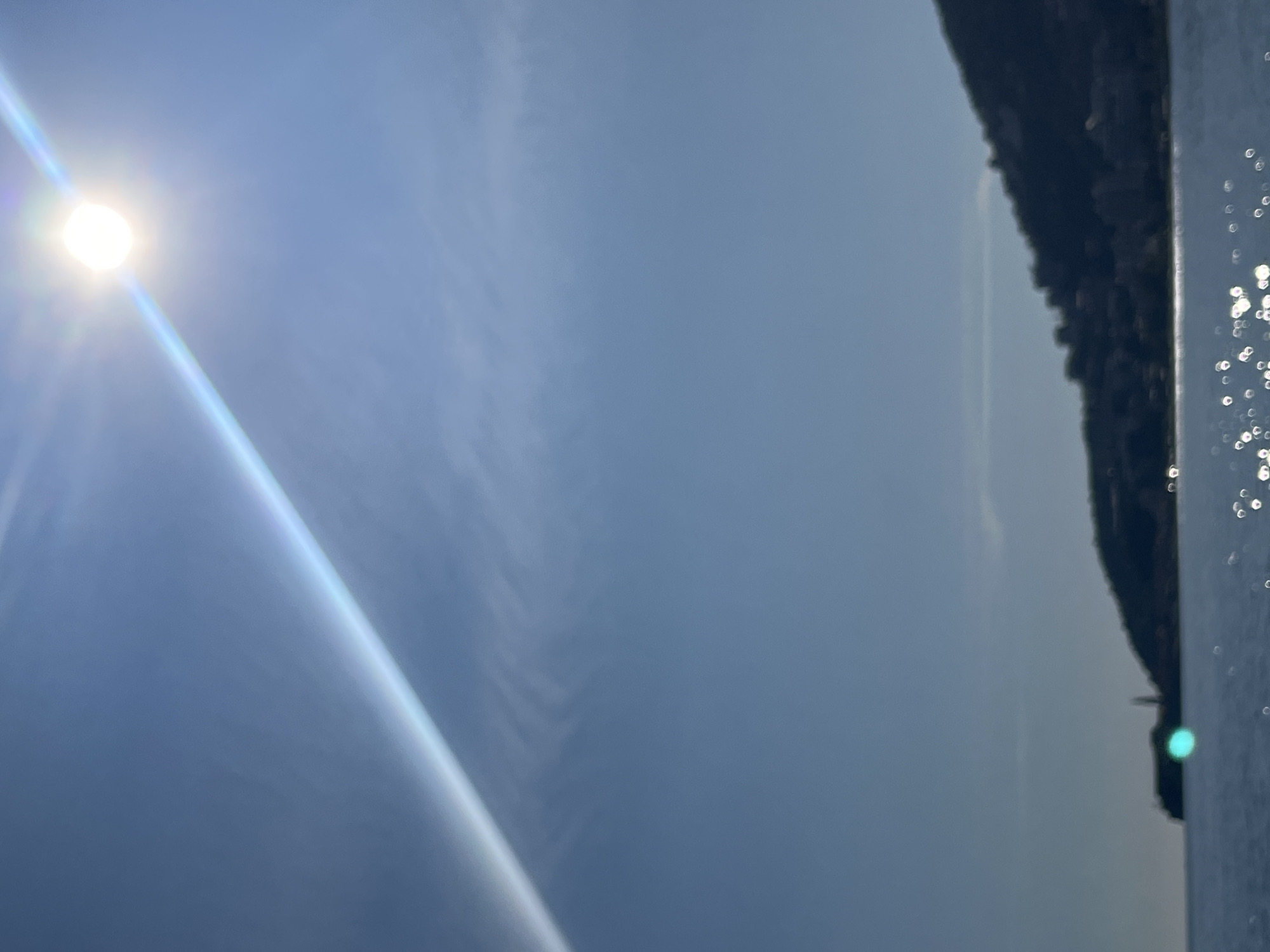 The image shows a daytime view in Budva, Montenegro. The sun is prominently visible in the upper left corner of the frame. The sky is predominantly blue, with subtle cloud formations stretching across the middle portion of the image. On the right side of the image, a dark, mountainous shoreline can be seen meeting the water. Small glistening points suggest sunlight reflecting off the water surface.