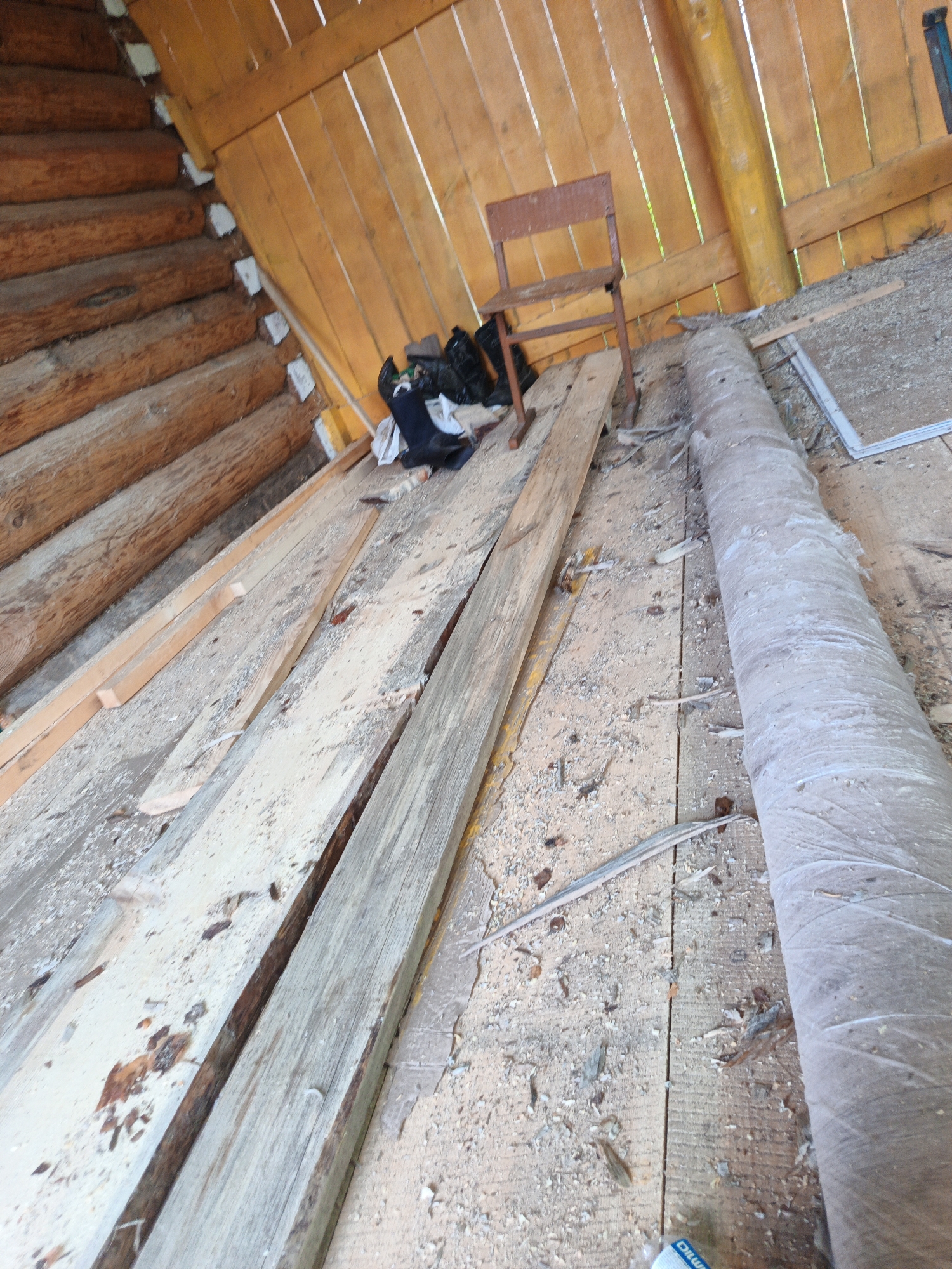 The image depicts the interior of a rustic wooden structure, likely an attic or shed, in the village of Islambaevo, Russia. The scene is cluttered with various wooden elements and debris. On the left, stacked logs form a section of the wall or floor, with rough-hewn planks laid horizontally. In the center, a simple wooden chair sits on the floor, accompanied by a pile of dark bags. Several long wooden planks are scattered across the floor, some appearing unfinished or weathered. A large, cylindrical object wrapped in gray material lies horizontally on the right side of the image, occupying a significant portion of the frame. The floor is covered in sawdust and wood chips, suggesting ongoing construction or renovation. The walls and ceiling are constructed from light-colored wooden planks, oriented vertically. The overall impression is one of a work-in-progress or a storage area in a rural setting.