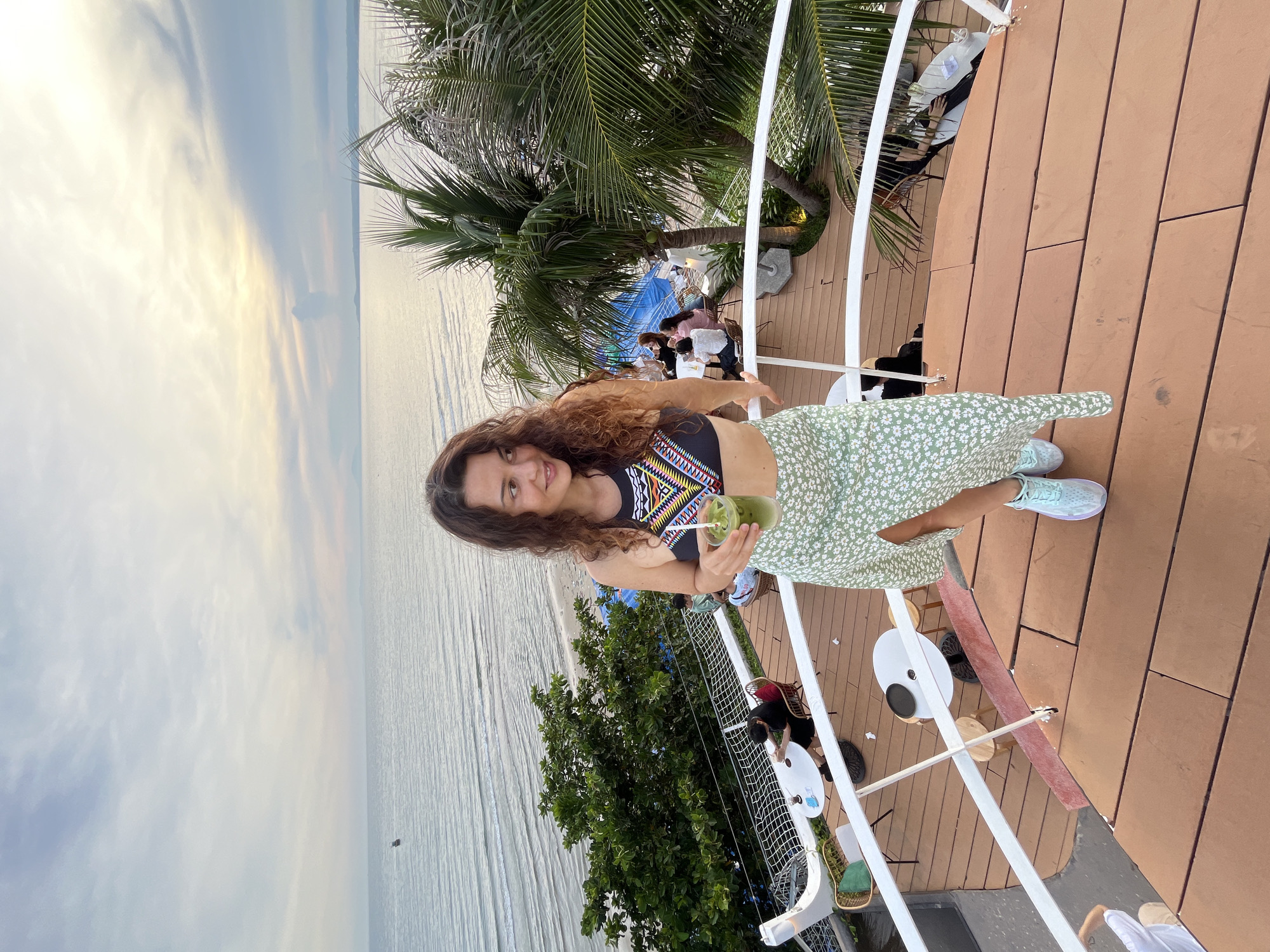 In Mũi Né, Vietnam, a woman with curly brown hair stands on a multi-level wooden deck overlooking the ocean at dusk. She is wearing a patterned halter top and a light green floral skirt, holding a glass of green liquid. Palm trees and lush greenery surround the deck, where other patrons are seated at tables. The sky is a gradient of soft blues and yellows, with the ocean stretching to the horizon. The setting appears to be a cafe or restaurant with an outdoor seating area.