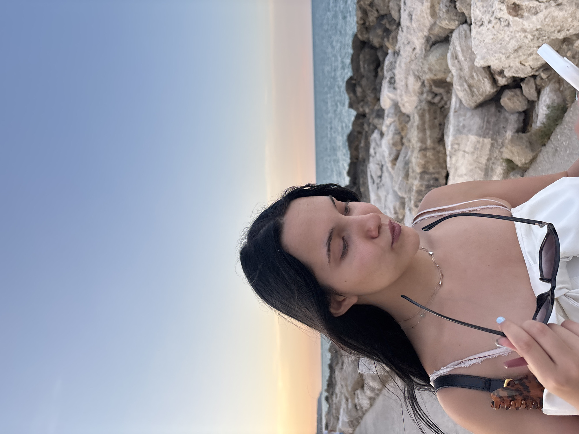 A young woman with long, dark hair stands outdoors, looking down and holding a pair of sunglasses. She is wearing a white top and necklaces. The background features a body of water, a rocky shore, and a clear sky with a gradient of blue and soft orange near the horizon. The scene appears to be a coastline, possibly along the Adriatic Sea in Grottammare, Italy, given the characteristic landscape of the area which often includes pebble beaches and seaside promenades. The lighting suggests it is late afternoon or early evening, with the sun low in the sky.