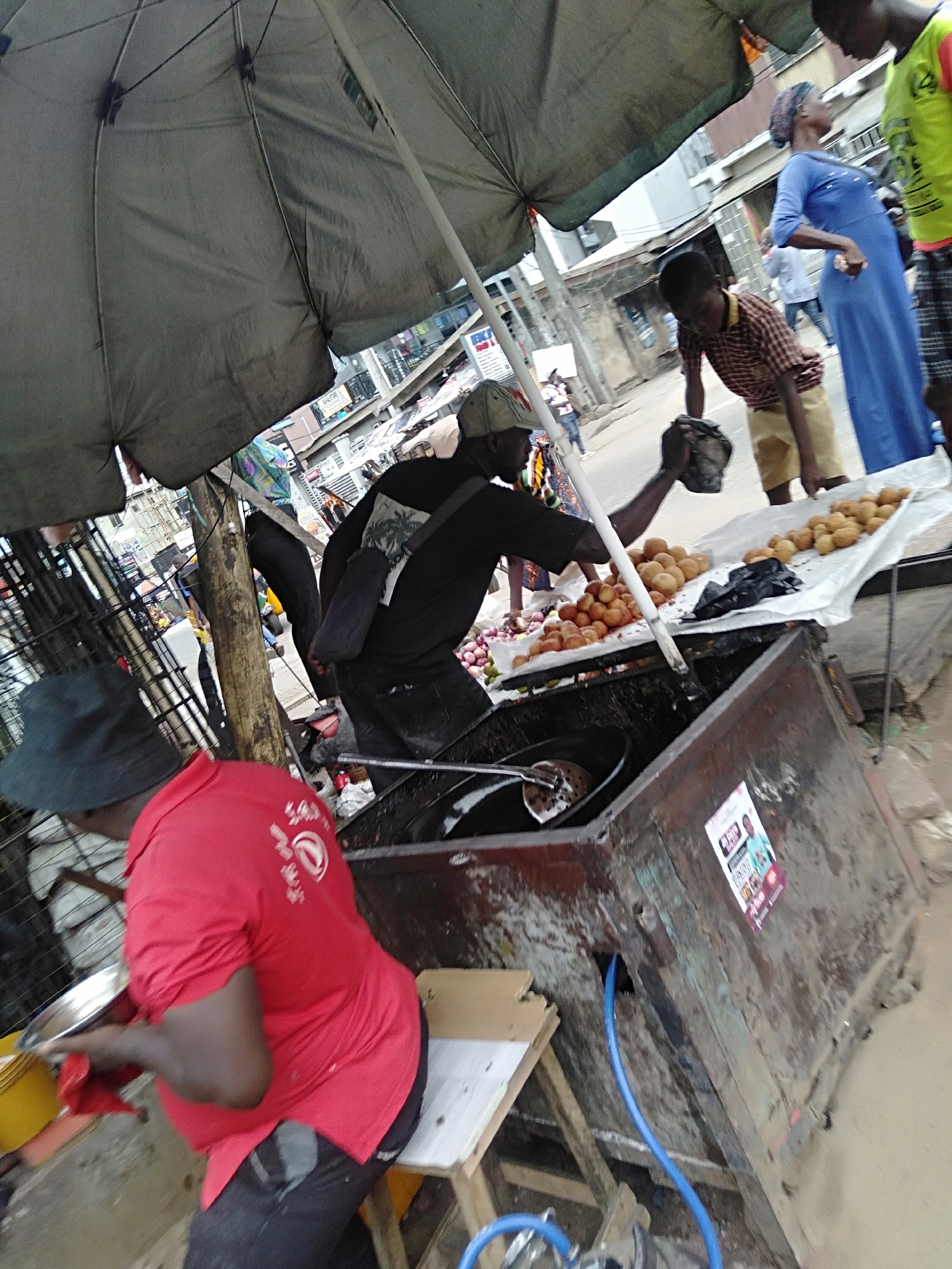 A street vendor in Shangisha, Nigeria, prepares and sells fried dough balls, possibly puff-puffs. The vendor in a red shirt and hat is seated on a small stool, working with a metal bowl and likely preparing the dough near a large, weathered metal cooking apparatus filled with frying oil. A second vendor, in a black shirt and cap, stands near a table laden with freshly fried dough balls and a plastic bag. Customers, including a child in a checkered shirt and an adult in a blue dress, are nearby. An umbrella provides shade. The scene takes place outdoors, on a street with other buildings and people visible in the background.