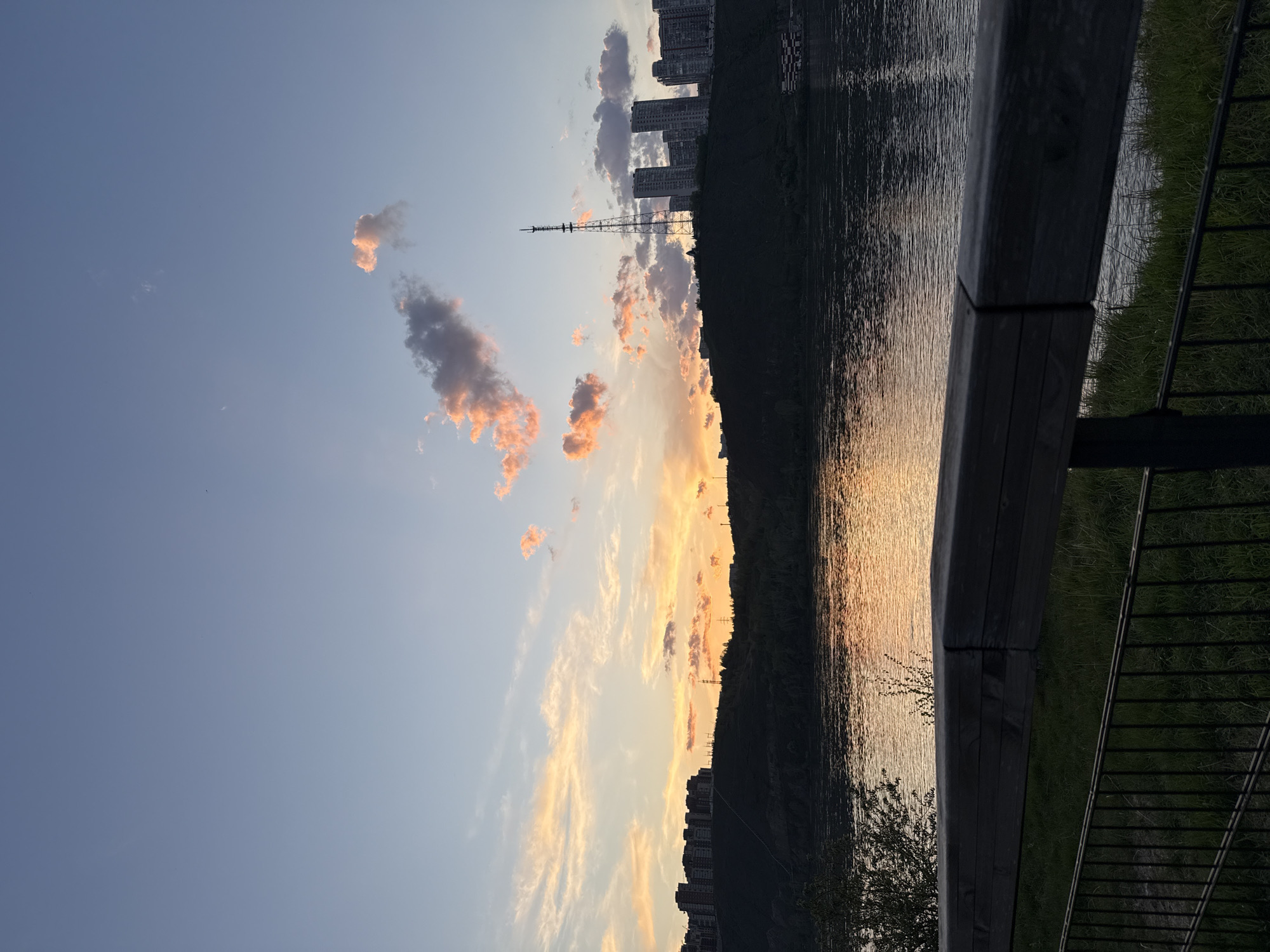 The image depicts a landscape scene at sunset or sunrise in Krasnoyarsk, Russia. The sky is a gradient of blue and orange/yellow, with scattered clouds tinged with pink and orange. The Yenisei River reflects the colors of the sky. A dark, tree-covered slope or hill runs along the right side of the image, partially obscuring a row of high-rise apartment buildings. A tall lattice tower, possibly a communication or transmission tower, is visible in the background. In the foreground, there is a wooden railing and metal fence, indicating a walkway or observation point. The scene is urban, with the riverfront developed.