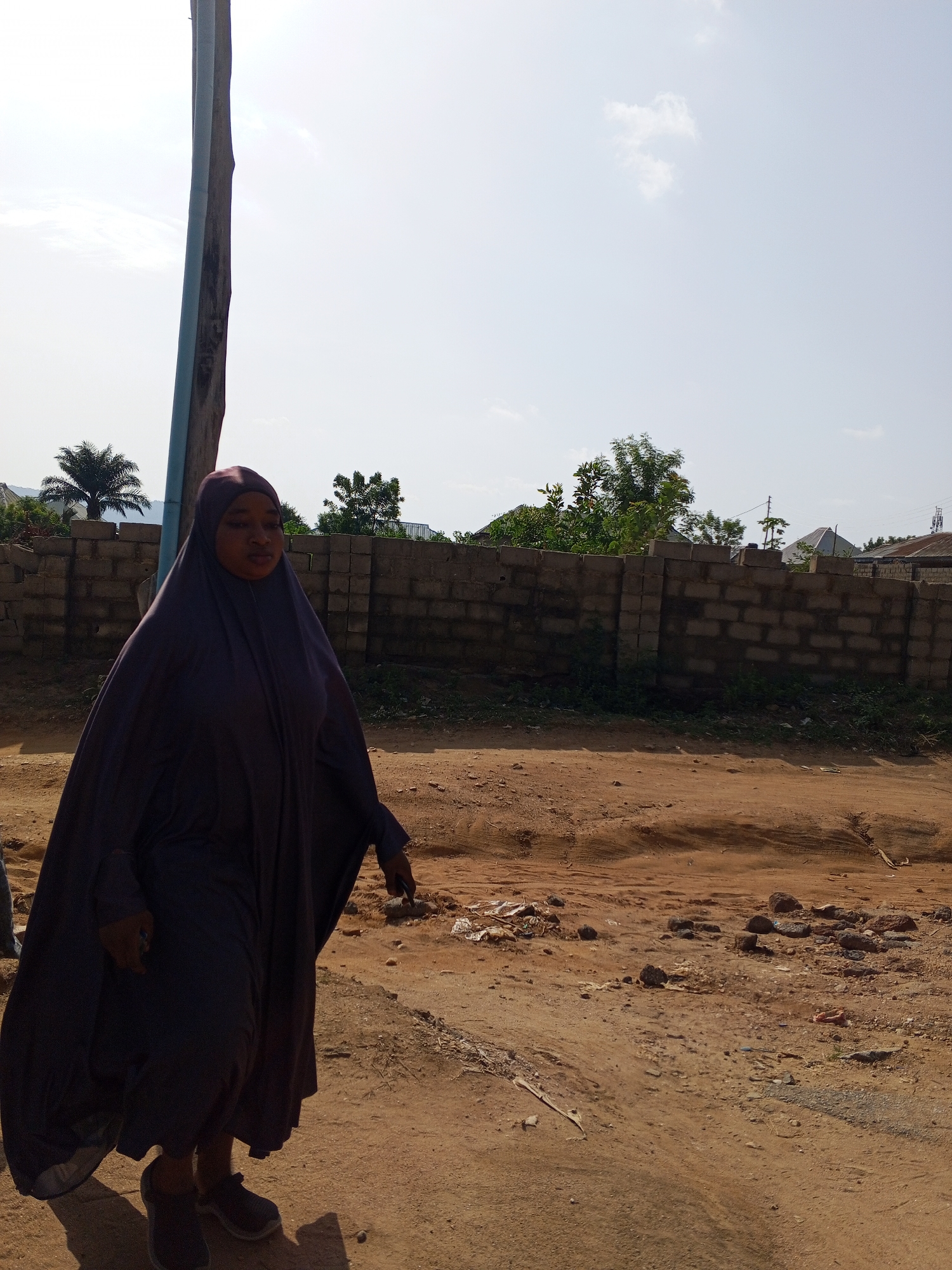 The image shows a woman walking or standing on an unpaved dirt path under a bright, sunny sky. She is the primary subject, positioned towards the left side of the frame, and is dressed in a dark purple or grey abaya/hijab that covers her head and body. She wears dark athletic shoes. Her face is visible but slightly obscured by shadows, and her expression is neutral.
To the left, a tall, slender pole, possibly a utility pole, with a blue section, extends vertically through the image. The ground consists of reddish-brown dirt, gravel, scattered rocks, and some small pieces of debris. In the mid-ground, a sturdy fence built from grey concrete blocks stretches horizontally, acting as a boundary. Behind this wall, various green trees, including a prominent palm tree on the far left, and residential structures with roofs are visible in the distance.
The scene depicts an outdoor, semi-urban or rural environment, consistent with areas like Adonkolo, Nigeria. The time of day is clearly daylight, with the presence of direct sunlight indicated by shadows and a bright, mostly clear sky with a few faint clouds. There is no visible text in the image.