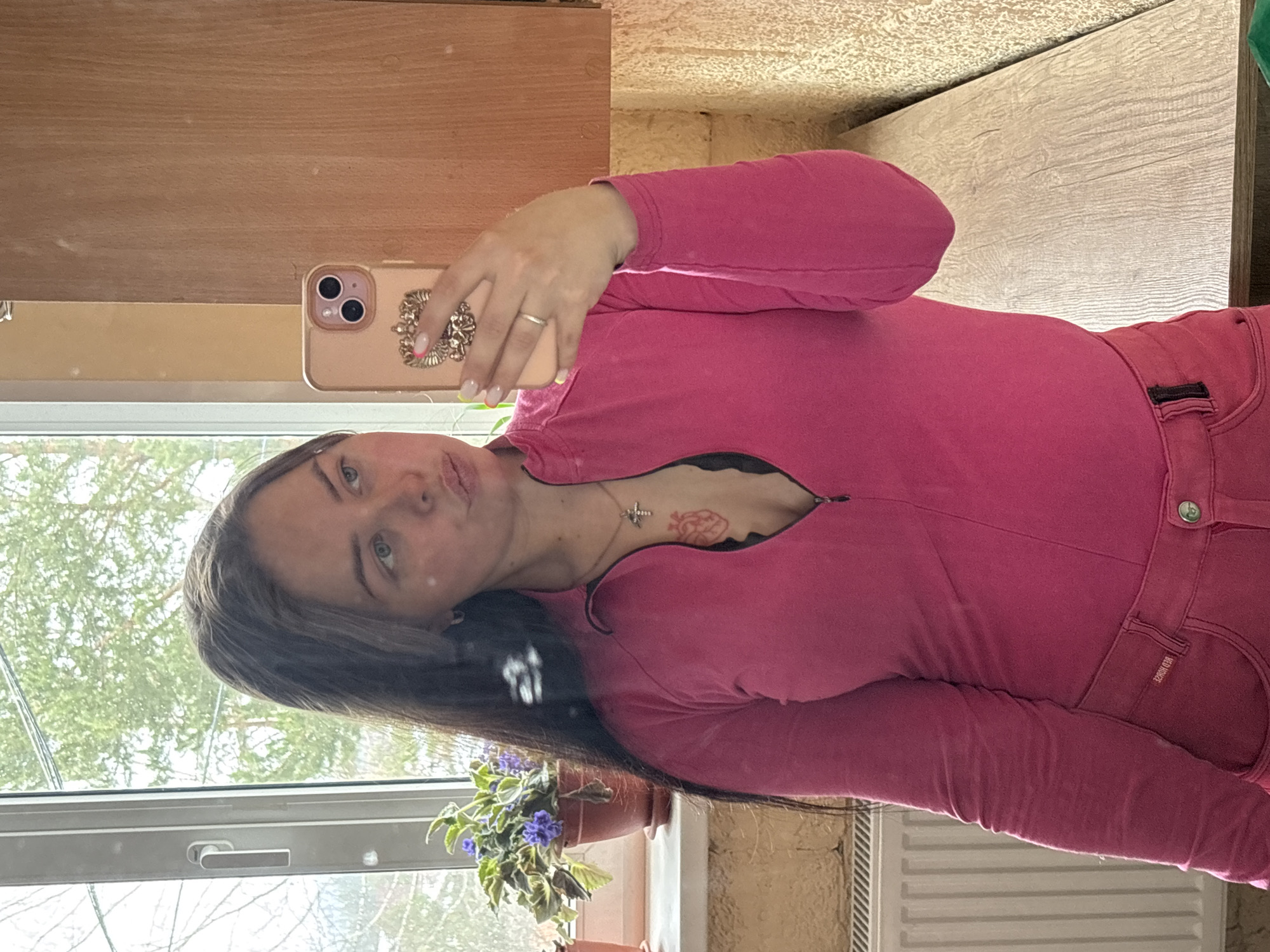 A woman in a pink zip-up jacket and pink jeans is taking a selfie in what appears to be a domestic interior, likely a house, based on the visible window and interior wall. The window looks out onto a leafy, partially obscured outdoor view. The scene is likely in Русский Вожой, Russia. A plant in a pot is on the windowsill. The woman is holding a smartphone with a decorative case. A cross and a heart tattoo are visible on her chest.