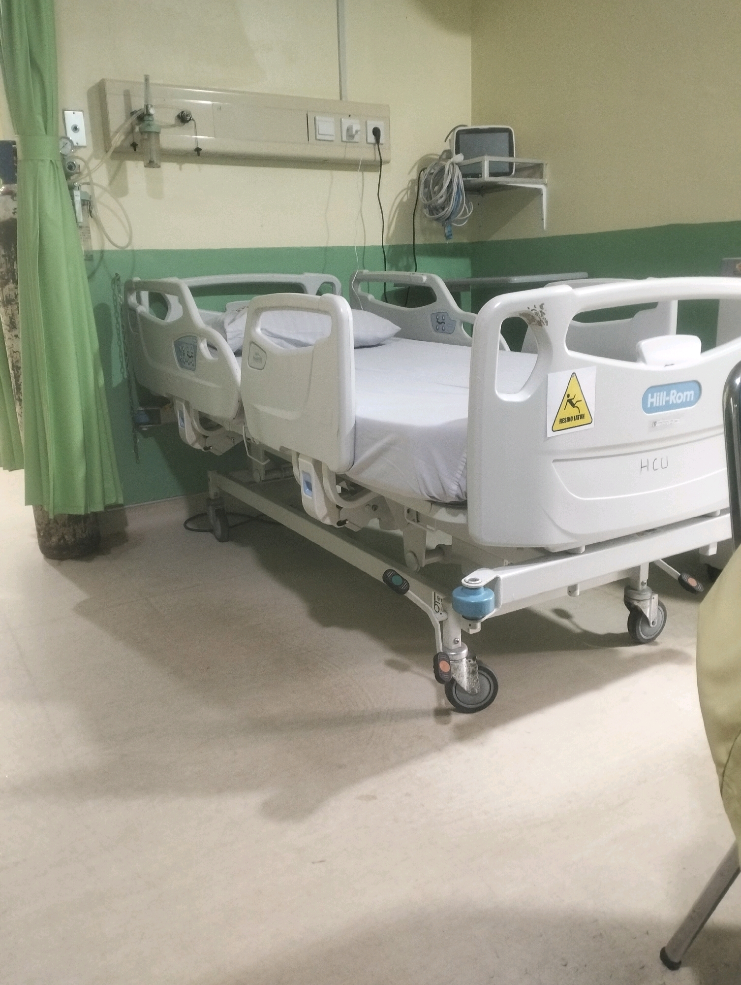 An empty hospital bed with a white mattress and gray sheets is shown in a room with light yellow walls and a green stripe running horizontally. The bed has a metal frame with wheels and various controls on the sides. A green curtain is visible to the left of the bed. Medical equipment, including a monitor on a shelf and a gas cylinder, are also present. A yellow warning sign with the words "RISIKO JATUH" (risk of falling) is attached to the bed's headboard. The floor is light-colored and reflects light. The room appears to be in a healthcare facility, possibly a hospital room. No people are visible in the image. The specific location in Air Jamban, Indonesia is not discernible from the image itself.