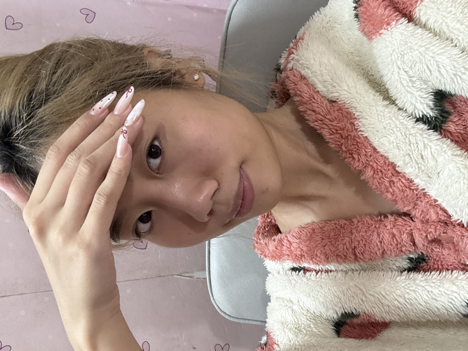 A woman poses in a sideways selfie. She's wearing a fuzzy, pink and white robe. Her hair is pulled back. One hand is raised near her forehead, showcasing long, almond-shaped nails with red ribbon designs. She has light skin, dark eyes, and a subtle smile. The background includes a pink patterned sheet and the edge of a light-colored chair.