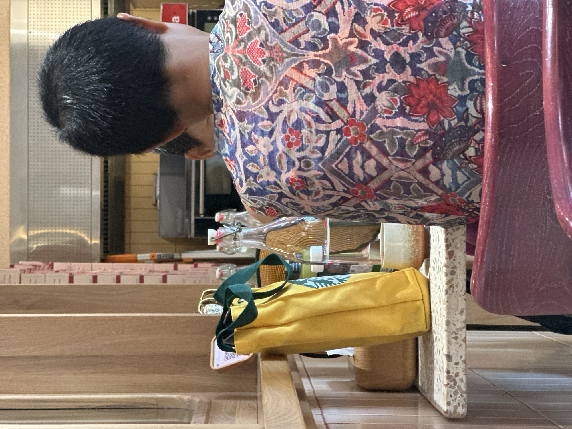 A rear view of a person wearing a patterned shirt with floral designs in shades of blue, purple, red, and pink is visible. They are seated at a table in what appears to be a cafe or restaurant. On the table, to the left, is a yellow tote bag with green straps, and next to it, a small white cup with an orange rim. Behind these items are several glass bottles. The background shows what seems to be a kitchen area with stainless steel appliances and shelving. The table has a speckled, terrazzo-like surface. The image is captured from a medium distance, focusing on the person and the items on the table.