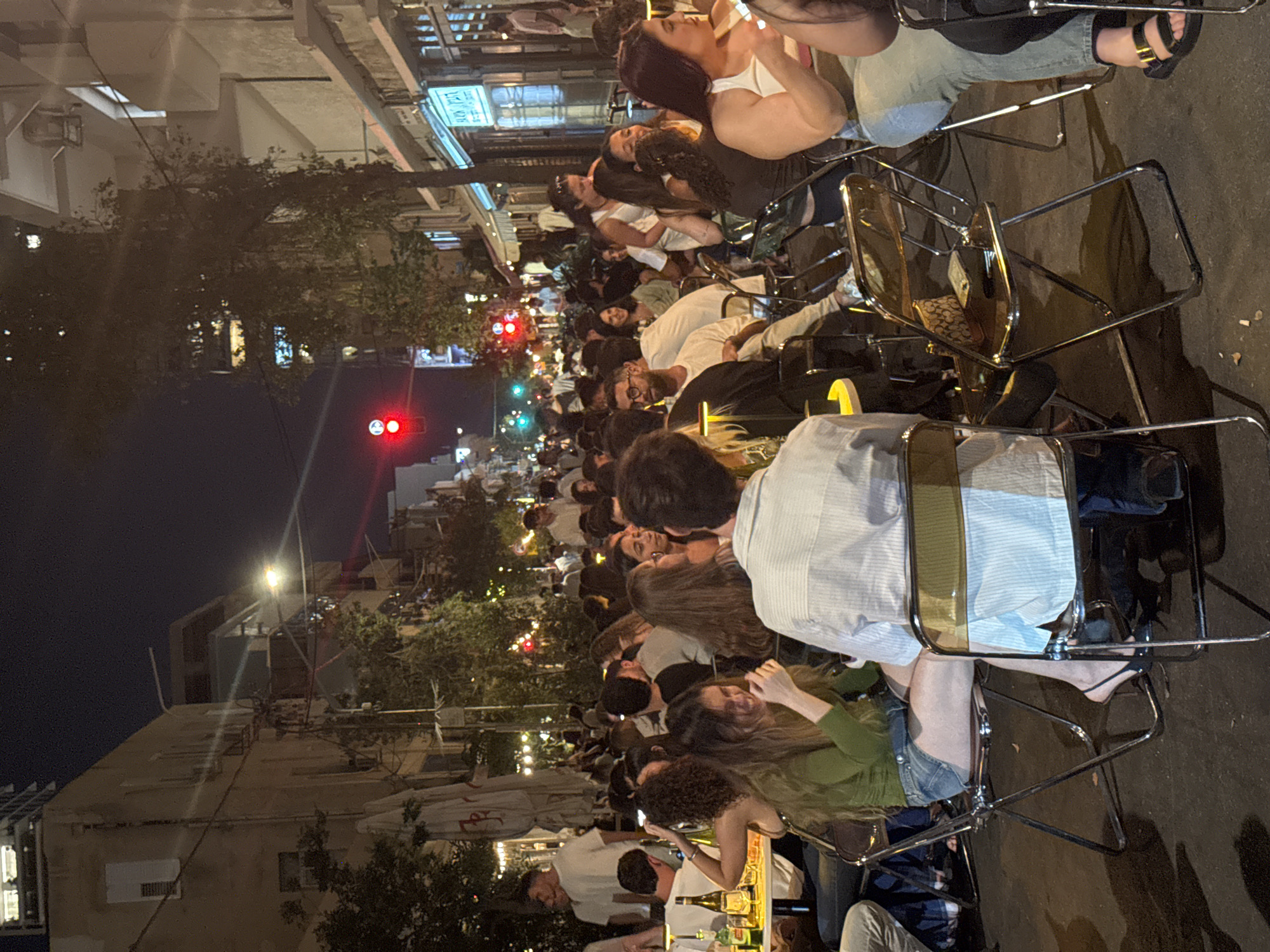 This image captures a vibrant nighttime street scene in Tel Aviv, Israel, depicting an outdoor dining and socializing area. Numerous individuals, primarily young adults, are seated at small, round tables arranged directly on the pavement. The tables are accompanied by distinctive folding chairs featuring chrome frames and translucent amber-colored plastic seats.
People are engaged in various social interactions, including conversing, laughing, and looking at their phones. In the foreground, a woman wearing a green long-sleeved top and denim shorts is seen laughing while seated at a table. Beside her, a man in a light blue striped shirt is seated facing away from the camera. On the far right, a woman in a white tank top and light-colored jeans holds a phone. Drinks, including bottles and glasses, are visible on multiple tables.
The urban environment is defined by multi-story buildings lining the street, some with balconies. Overhead power lines are visible against the dark sky. Trees with dense foliage are interspersed along the street, providing some natural elements. Storefronts on the right side of the street display illuminated signs with Hebrew text, confirming the location as Tel Aviv. One prominent sign is blue with white Hebrew lettering. A large, closed beige outdoor umbrella stands on the left.
Artificial lighting from streetlights, interior building lights, and ambient glow illuminates the scene. Two traffic lights are visible further down the street, one red and one green. The ground is a dark paved surface. The overall atmosphere is bustling and social, indicative of a popular nightlife district.