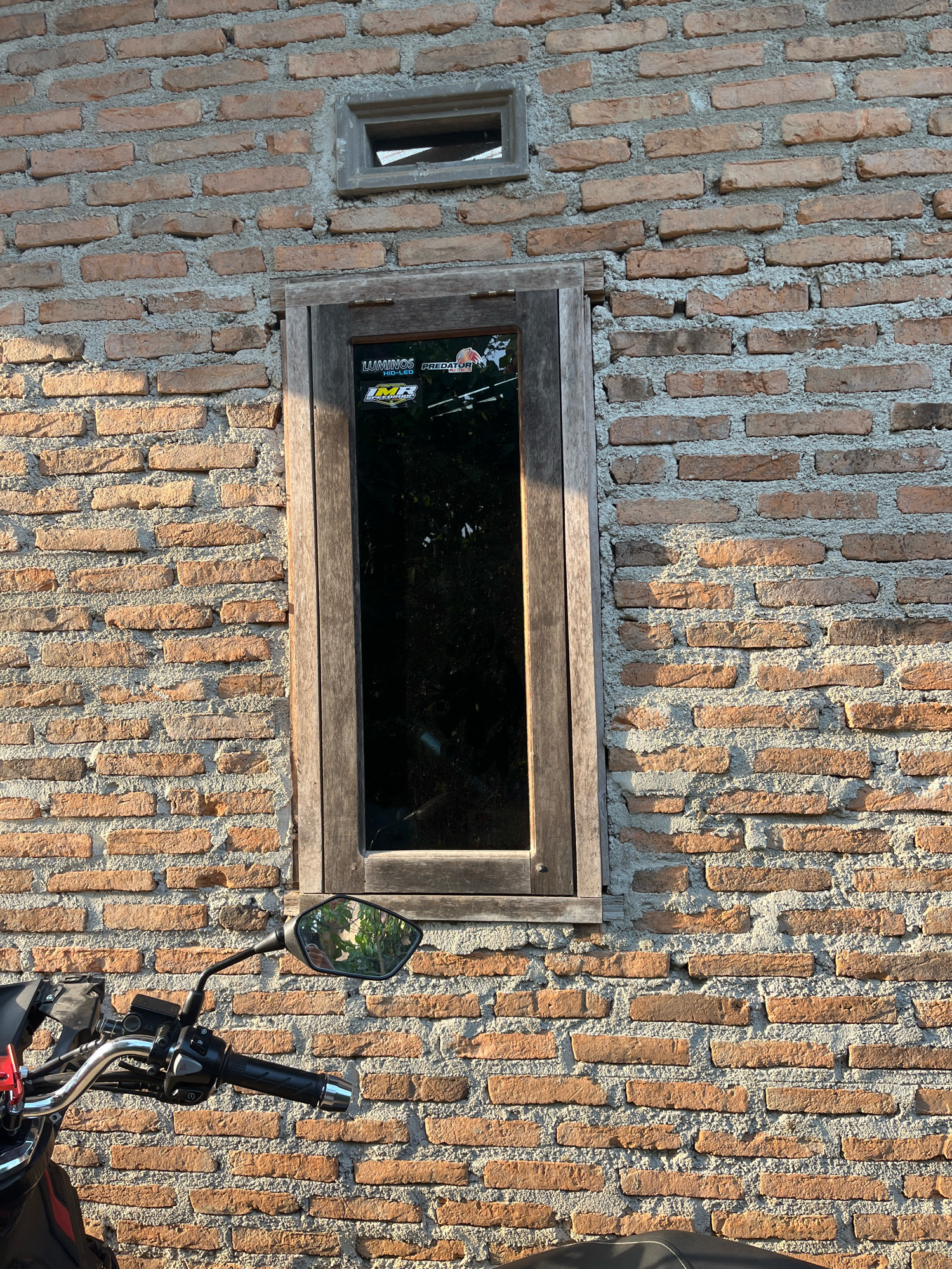 In Kebumen, Indonesia, a brick wall serves as the backdrop for a motorcycle and a wooden-framed window. The wall, constructed from reddish-brown bricks and grey mortar, exhibits a rough texture. Sunlight casts shadows across the uneven surface, highlighting the construction.
Above the motorcycle's handlebars, a rear-view mirror reflects a glimpse of foliage, suggesting an outdoor setting. The motorcycle itself is partially visible, with its handlebars, control levers, and part of the seat in the frame. The wooden window, positioned above the motorcycle, has a dark pane, obscuring any view beyond. A smaller, rectangular vent is situated above the window. The overall scene suggests a quiet, possibly residential or commercial area in Kebumen, Indonesia.