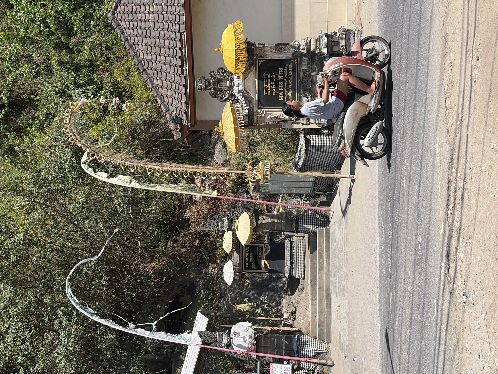 A person rides a scooter down a paved road in front of a temple entrance on Nusa Penida, Indonesia. The temple entrance features stone pillars adorned with black and white checkered fabric. Above the pillars are large decorative banners, one white and one yellow and green, with decorative elements resembling palm fronds. Several white and yellow umbrellas are strategically placed around the entrance. A signpost reads "GOA GIRI PUTRI". The temple is situated on a slight incline, with steps leading up to it. Lush green foliage covers the hillside behind the temple. The sky is bright blue and clear.