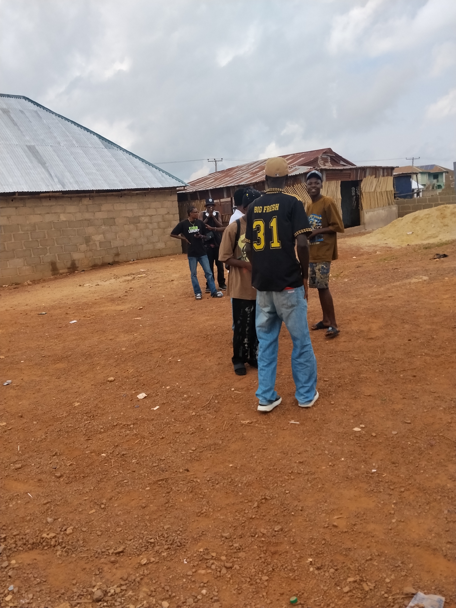 In the city of Durumi, Nigeria, a group of young men stand on a dirt road in front of a building with a corrugated metal roof. The man in the foreground wears a black t-shirt with "BIG FRESH 31" emblazoned on the back, paired with blue jeans and white sneakers. To his right, a man in a tan t-shirt and patterned shorts smiles, and another man in a tan t-shirt and black pants stands beside him. Behind them, a brick building with a corrugated metal roof stretches across the background. The ground is covered in reddish-brown dirt, with scattered debris, suggesting an outdoor, possibly developing area. The sky above is overcast.