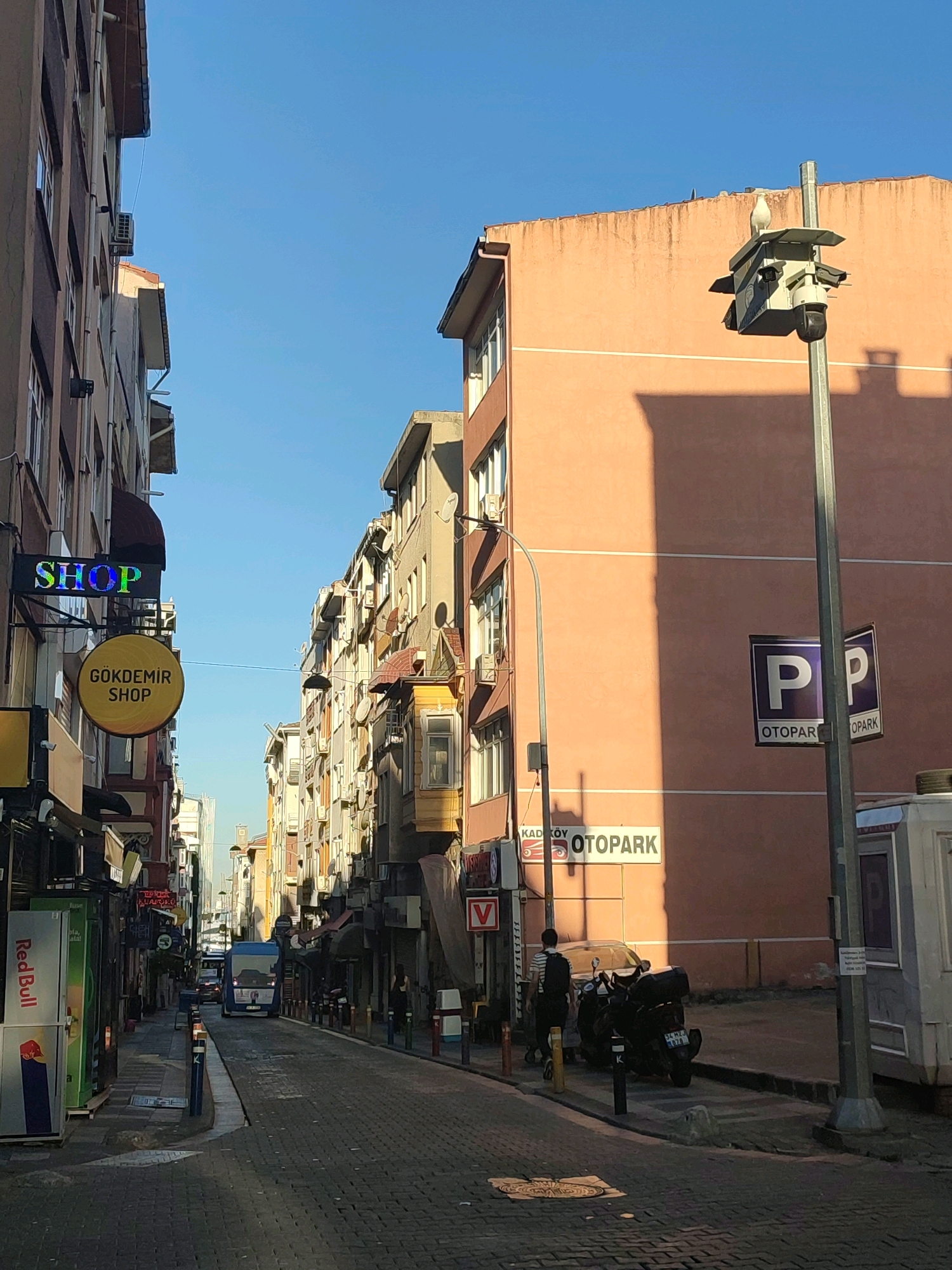 The image captures a street scene in Kadıköy, Turkey, characterized by its narrow, cobblestone pavement and tightly packed, multi-story buildings. The architecture suggests a dense urban environment, likely a commercial or residential area. On the left side of the street, a shop with a neon "SHOP" sign is visible, along with a circular yellow sign that reads "GÖKDEMİR SHOP". Further down, a Red Bull vending machine stands outside a shop. The buildings on both sides of the street are adorned with numerous windows, air conditioning units, and various advertisements. A blue bus is seen in the distance, navigating the street.
On the right side of the image, a large, peach-colored building dominates the foreground. A prominent street lamp stands tall, topped with a birdhouse-like structure that a white bird is perched on. Two purple and white "OTOPARK" (parking) signs are affixed to the lamp post, indicating a parking area. Below these signs, a sign with the text "KADIKÖY OTOPARK" is also visible. In front of the peach building, a parked black motorcycle and a person walking away from it are present. A white utility vehicle or building component is situated to the right of the lamp post. The overall lighting suggests it is daytime, with sunlight casting shadows across the scene. The sky is clear and blue.