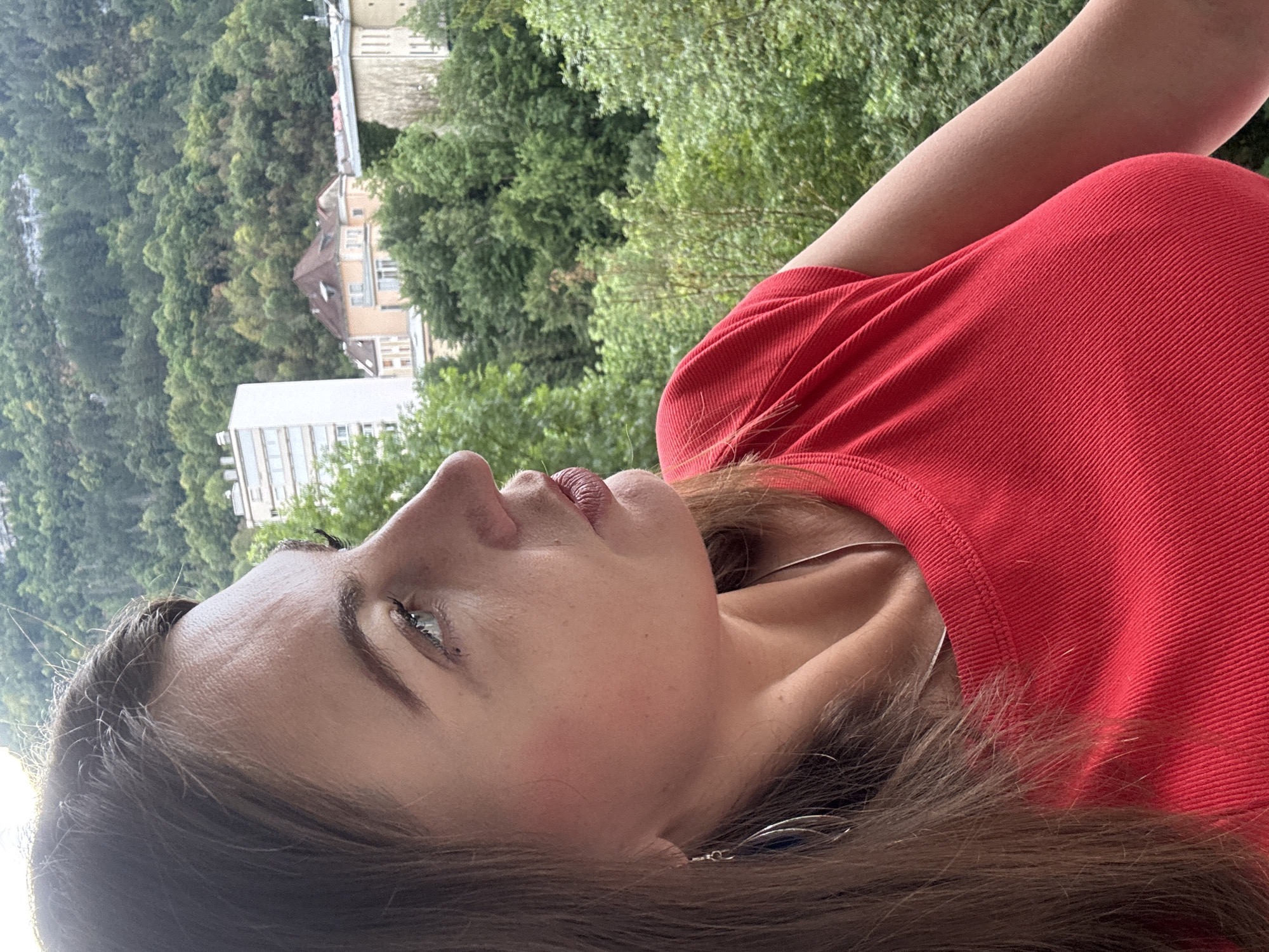 A medium shot captures a woman in profile against a backdrop of lush green trees and buildings in Saint-Claude, France. The woman, positioned on the left of the frame, has long brown hair that falls over her shoulders. She is wearing a bright red, ribbed top with a necklace visible at her collarbone. A delicate silver earring with a dark blue pendant dangles from her left ear. Her gaze is directed off to the right, and her expression is neutral. The background reveals a town nestled amidst a hilly, tree-covered landscape, with a tall, pale building and other structures visible. The lighting suggests it is daytime.