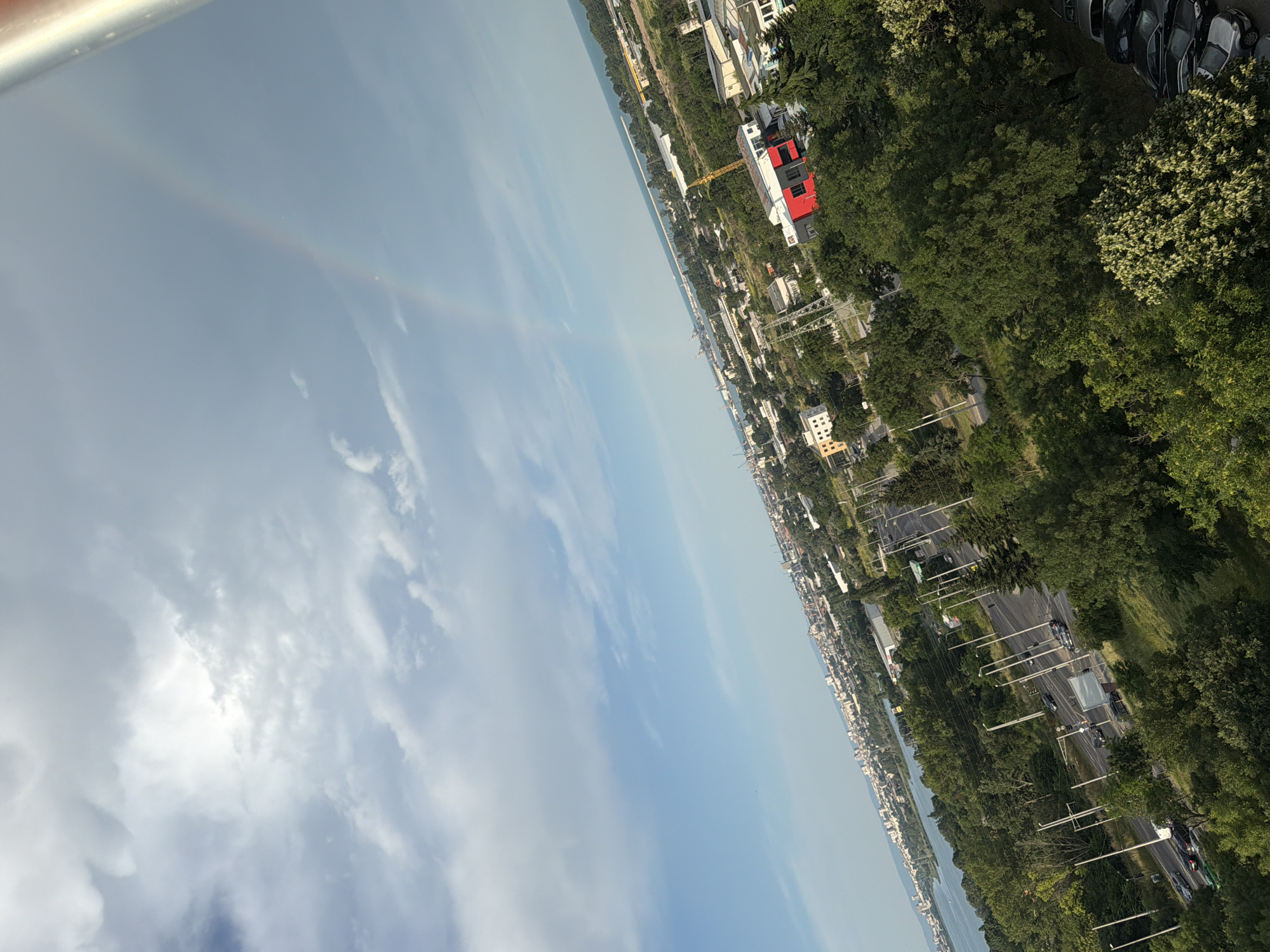 This image, taken from a high vantage point in Burgas, Bulgaria, captures a panoramic view of the city, including its port and the Black Sea. The sky is partly cloudy with a visible rainbow arcing across it. Below the sky, a sprawling cityscape unfolds, characterized by numerous buildings of varying sizes and architectural styles, intermingled with lush green trees. A prominent red and white building stands out in the foreground, possibly a commercial or industrial structure. Roads with vehicles can be seen, suggesting ongoing urban activity. The port area is visible in the distance, with cranes and ships indicating maritime operations. The overall scene depicts a vibrant coastal city with a blend of urban development and natural greenery, under a dramatic sky.